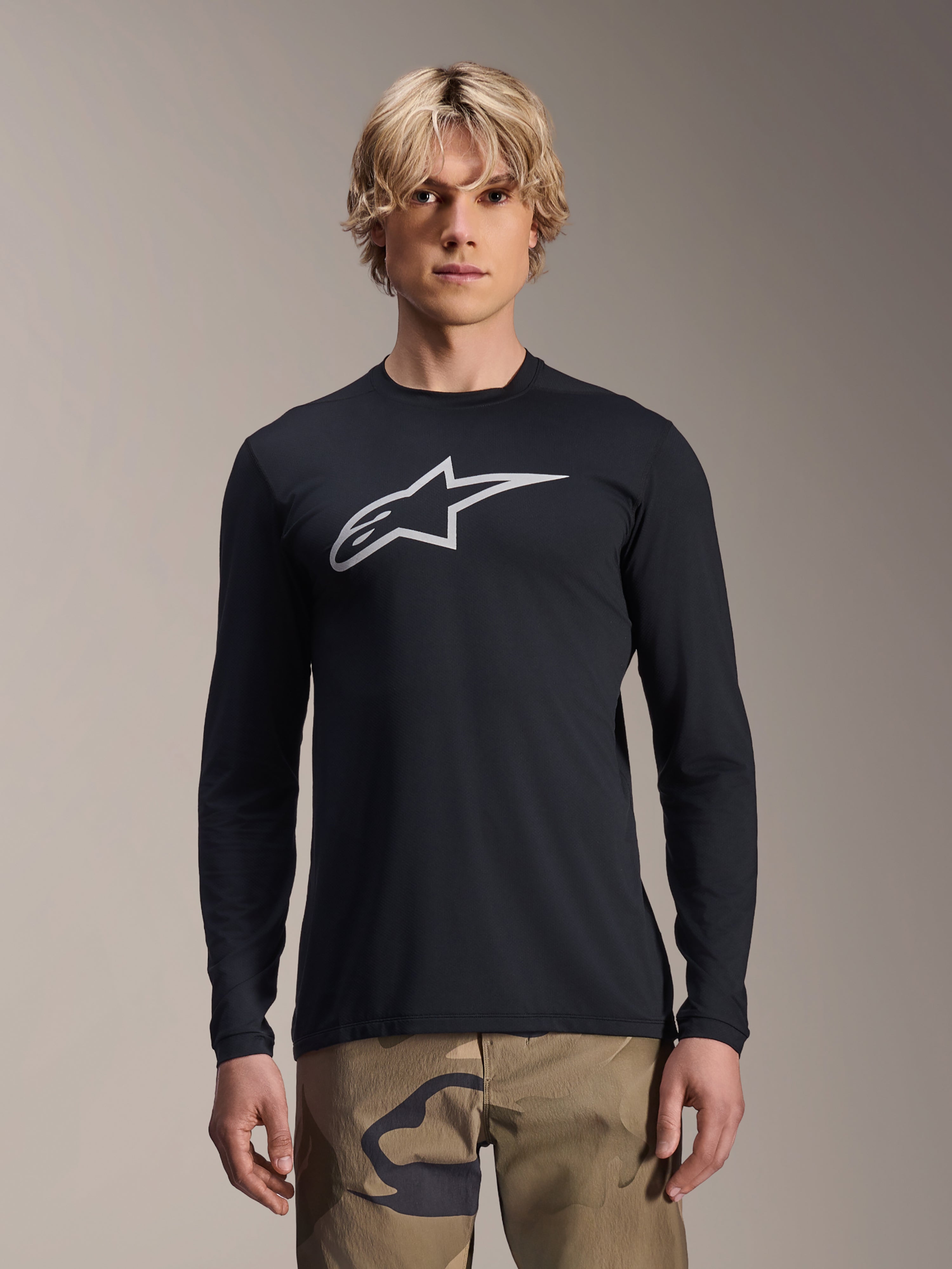 Alpinestars A-Dura Astar Jersey met lange mouwen, zwart met zilveren Astars-logo op de borst, gedragen door een model met camouflagebroek, casual MTB-performancekleding voor mountainbiken