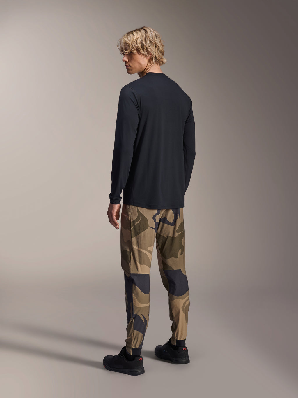 Alpinestars A-Dura Camo broek, MTB-broek, bruine camouflage, gedragen met een zwart jersey met lange mouwen en zwarte sneakers, achteraanzicht met toelopende pijpen en elastische manchetten, prestatiegerichte kleding voor off-road fietsen