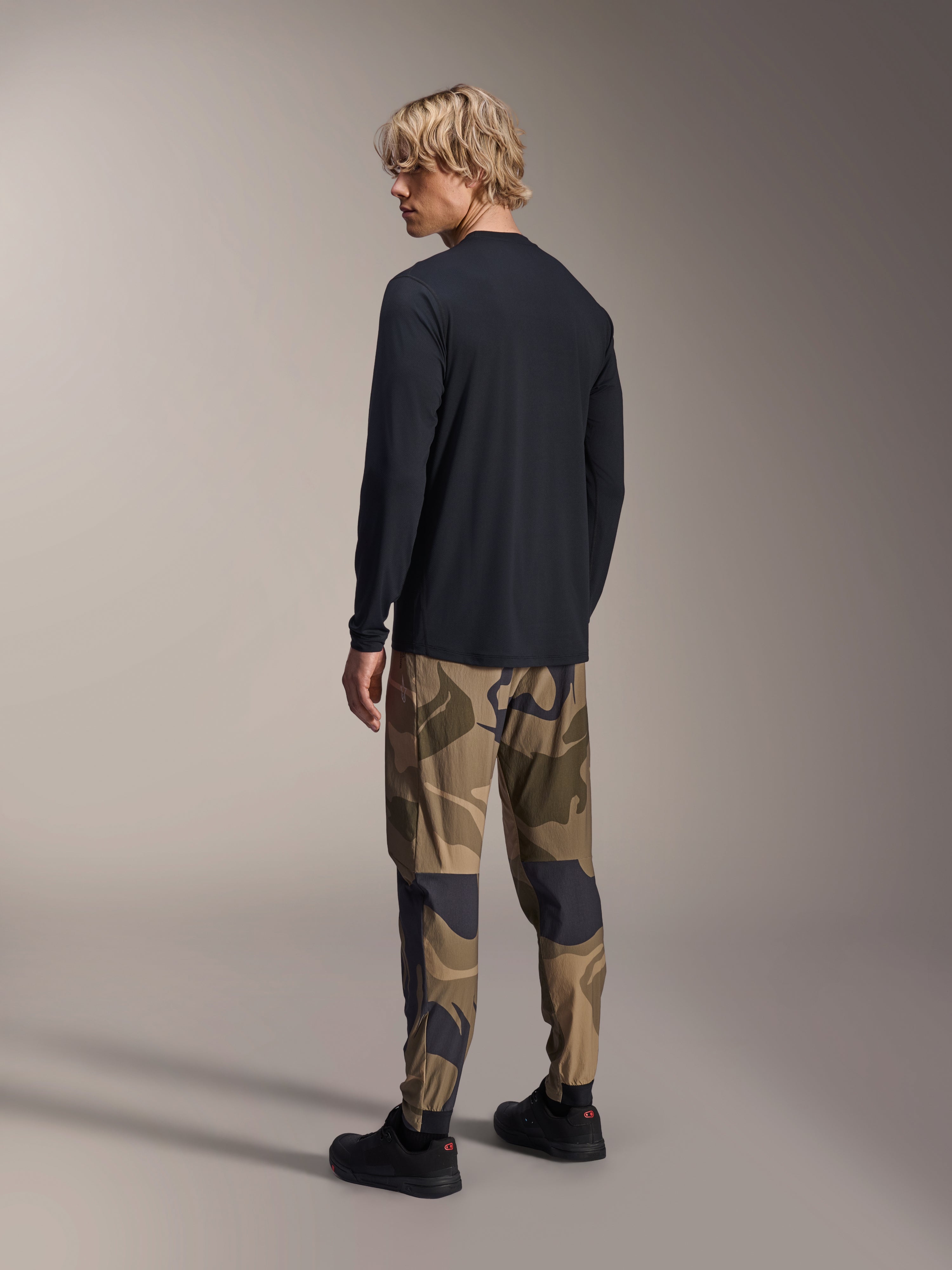 Alpinestars A-Dura Camo broek, MTB-broek, bruine camouflage, gedragen met een zwart jersey met lange mouwen en zwarte sneakers, achteraanzicht met toelopende pijpen en elastische manchetten, prestatiegerichte kleding voor off-road fietsen