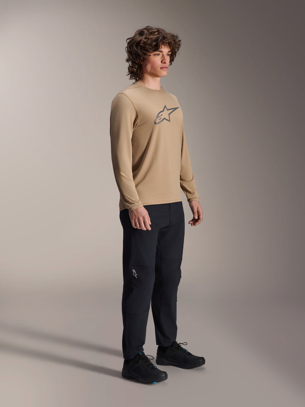 Alpinestars A-Dura Astar Jersey met lange mouwen, lichtbruin, gedragen door een model met een zwarte broek en schoenen, voorzien van een grijs Astars-logo op de borst, casual mountainbike performance gear