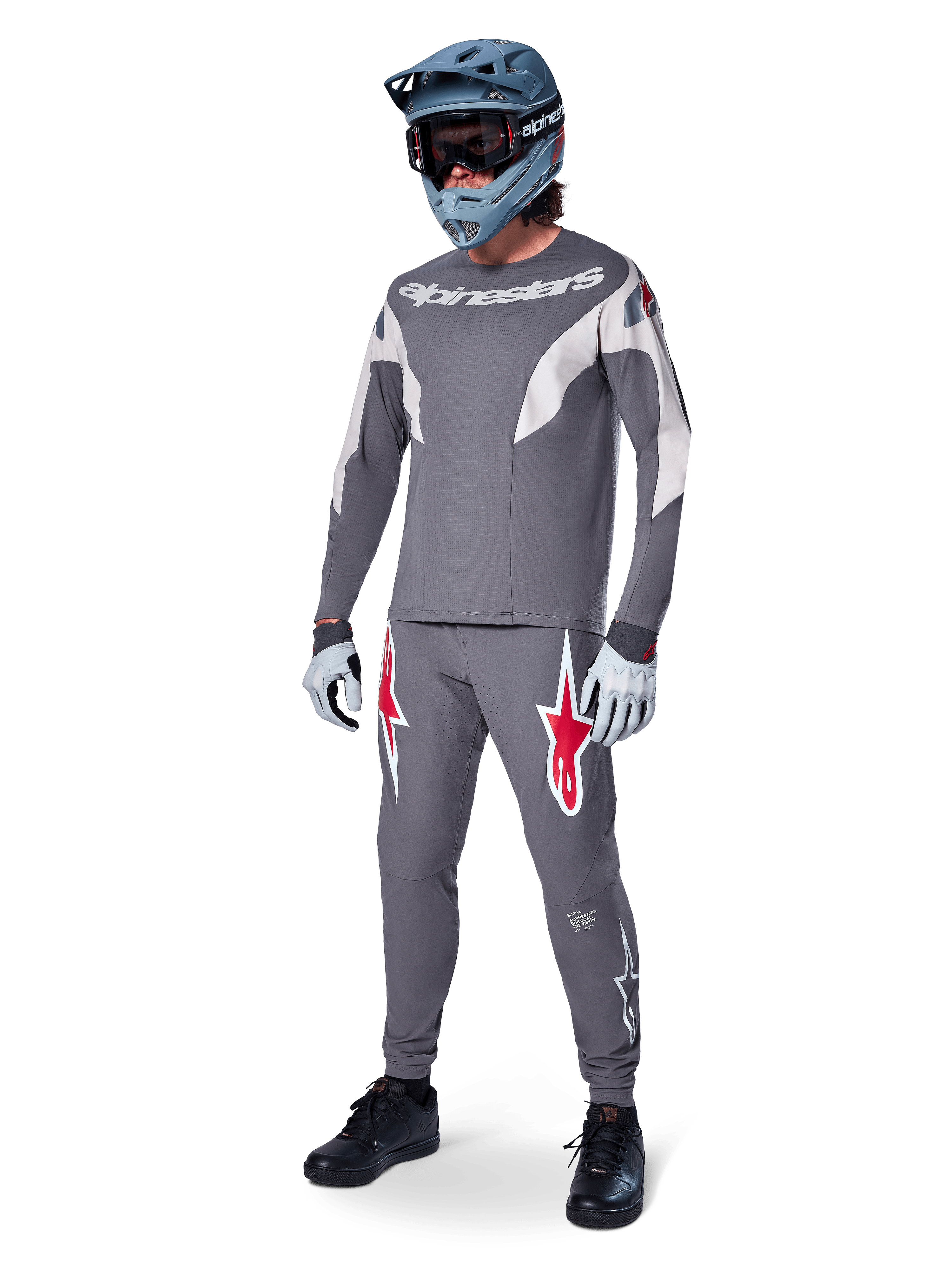 MTB rider wearing Alpinestars A-Supra Race Jersey, Long Sleeve, Dark Gray with light gray accents, matching dark gray pants featuring red star logo, gray full-face helmet with goggles, light gray gloves, coordinated outfit for mountain bike racing
