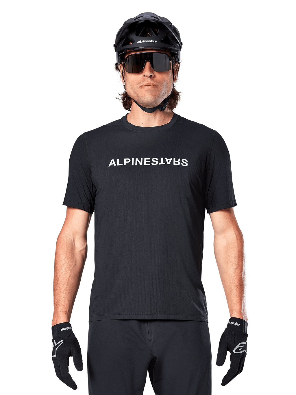Mountain bike rider wearing the Alpinestars A-Dura Switch Short Sleeve Jersey in Black, featuring a white wordmark logo on the chest, styled with a black helmet, large sunglasses, and black gloves, professional cycling apparel for trail riding.