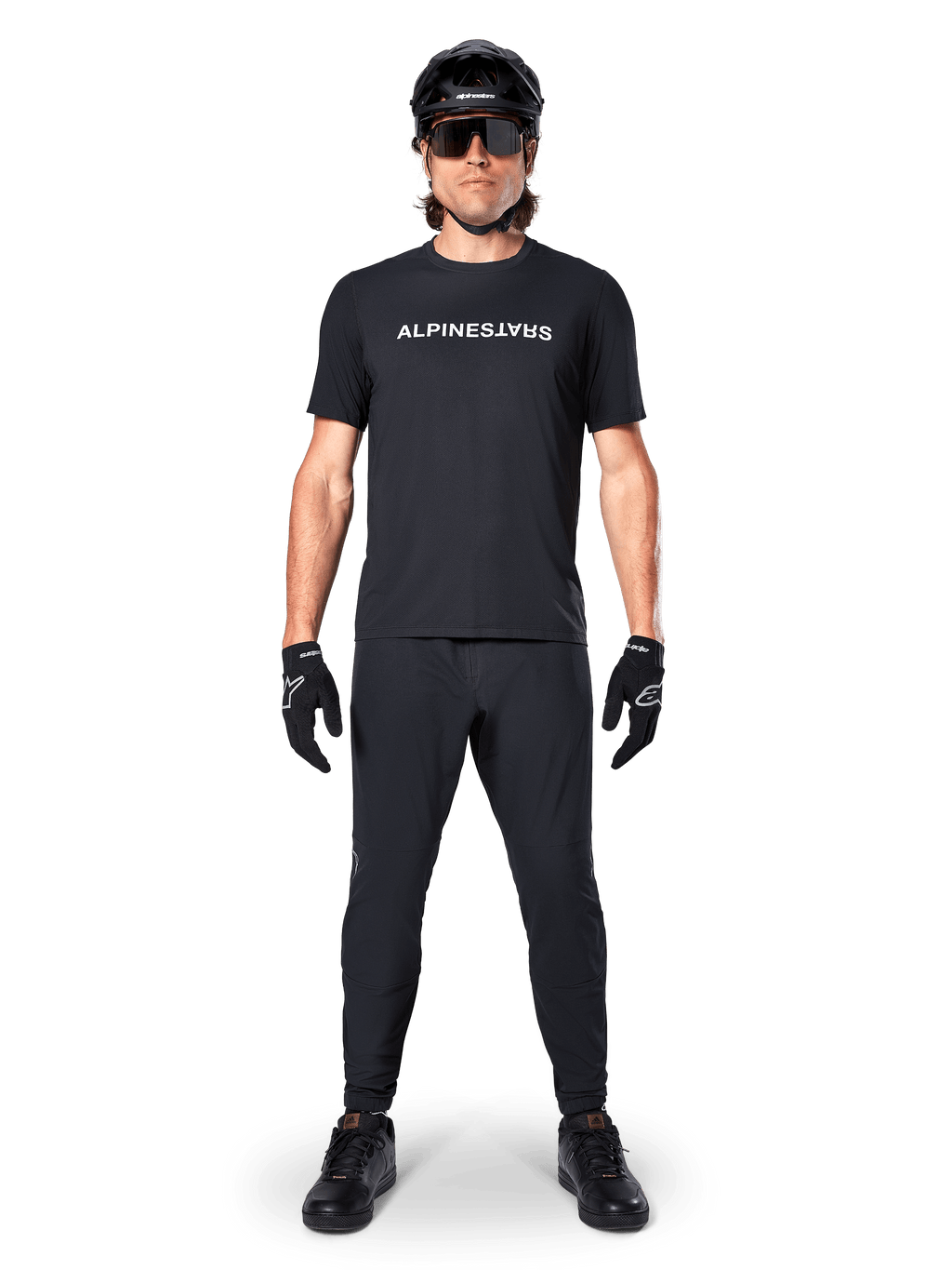 Mountain bike rider wearing the Alpinestars A-Dura Switch Short Sleeve Jersey in Black, featuring a white stylized wordmark logo on the chest, paired with matching black MTB pants, helmet, and gloves, complete trail-ready cycling outfit.