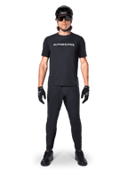 Mountain bike rider wearing the Alpinestars A-Dura Switch Short Sleeve Jersey in Black, featuring a white stylized wordmark logo on the chest, paired with matching black MTB pants, helmet, and gloves, complete trail-ready cycling outfit.