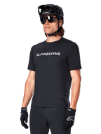 Alpinestars A-Dura Switch Short Sleeve Jersey, Black, featuring a white wordmark logo on the chest, worn by a cyclist with a black MTB helmet and sunglasses, technical apparel designed for trail riding