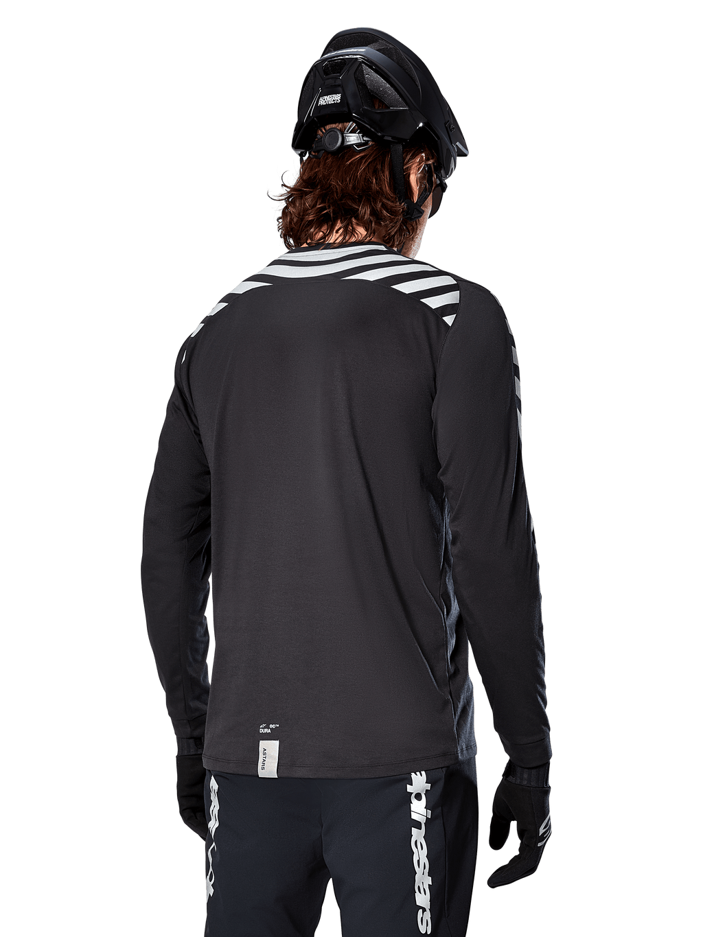 Alpinestars A-Dura Dri Oscar Jersey met lange mouwen, Black Reflective, achteraanzicht met reflecterende diagonale strepen op de schouders en een langere achterzijde, gedragen met zwarte MTB-helm en handschoenen, technische kleding voor mountainbiken