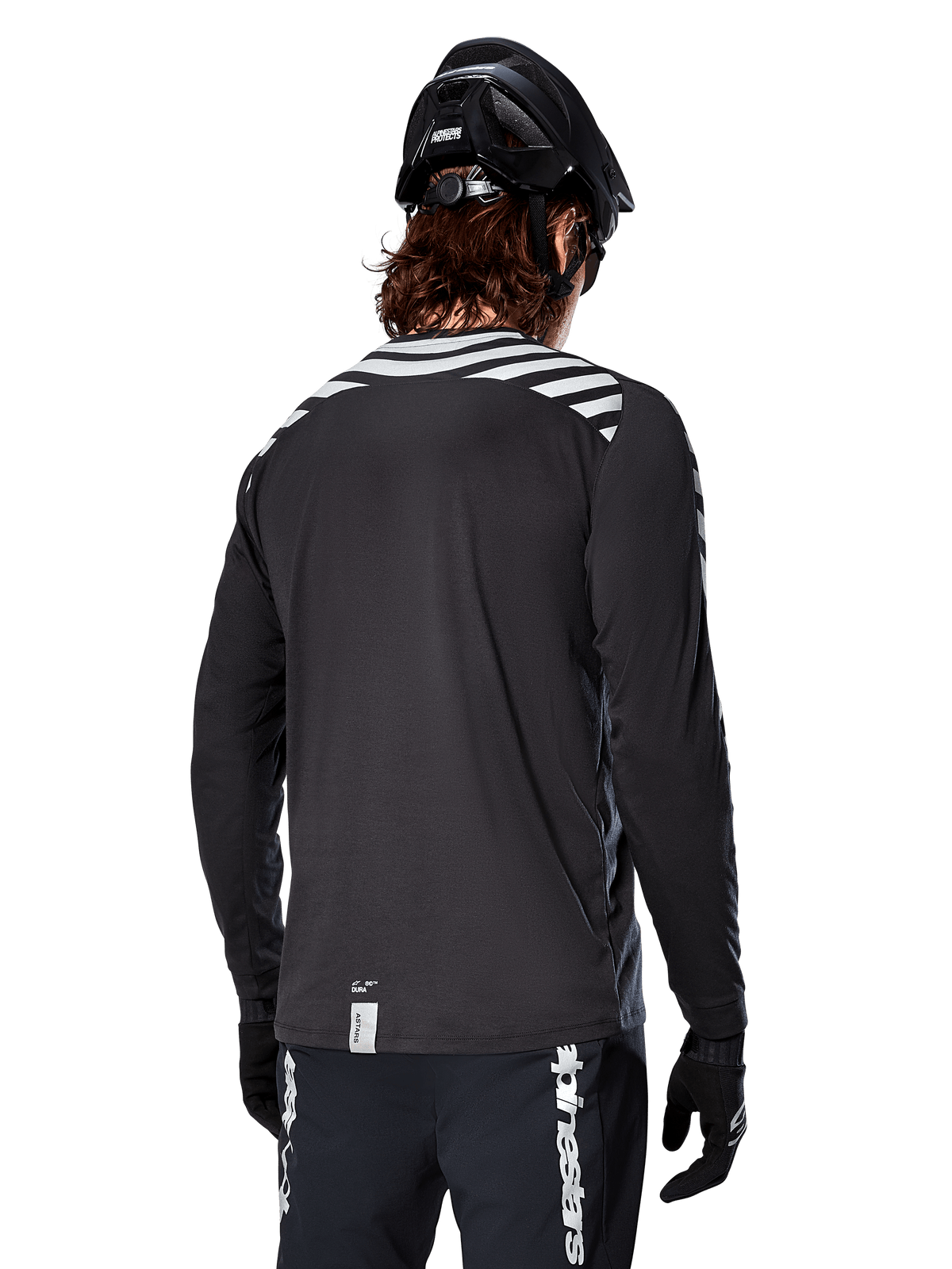 Alpinestars A-Dura Dri Oscar Jersey met lange mouwen, Black Reflective, achteraanzicht met reflecterende diagonale strepen op de schouders en een langere achterzijde, gedragen met zwarte MTB-helm en handschoenen, technische kleding voor mountainbiken