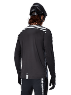 Alpinestars A-Dura Dri Oscar Jersey met lange mouwen, Black Reflective, achteraanzicht met reflecterende diagonale strepen op de schouders en een langere achterzijde, gedragen met zwarte MTB-helm en handschoenen, technische kleding voor mountainbiken
