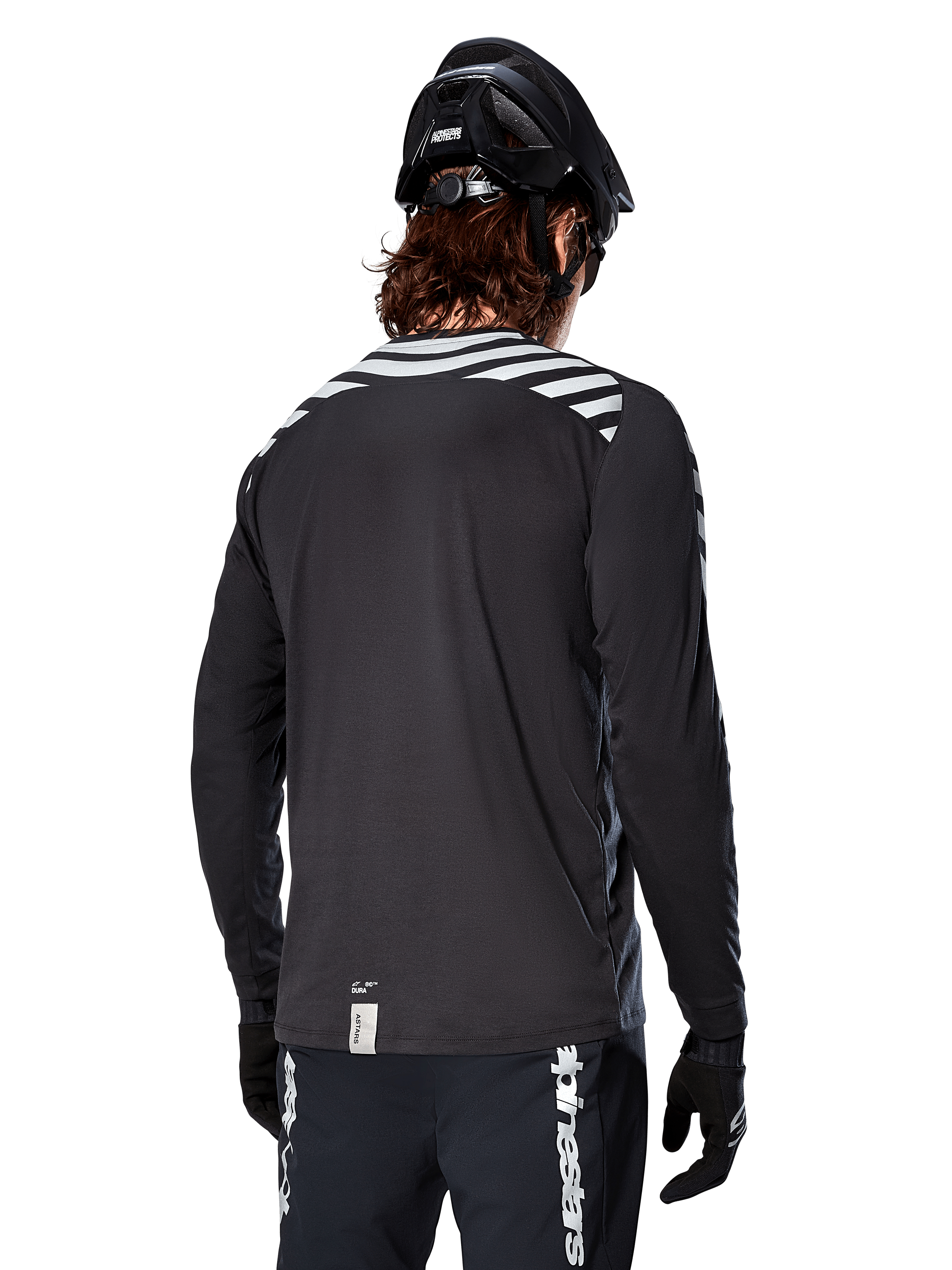 Alpinestars A-Dura Dri Oscar Jersey met lange mouwen, Black Reflective, achteraanzicht met reflecterende diagonale strepen op de schouders en een langere achterzijde, gedragen met zwarte MTB-helm en handschoenen, technische kleding voor mountainbiken