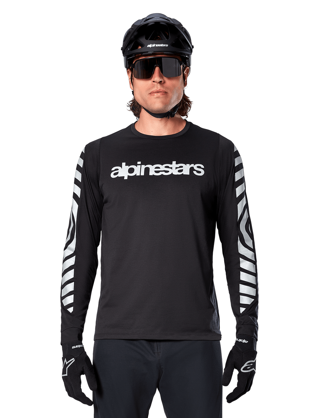 Mannelijke rijder in Alpinestars A-Dura Dri Oscar Jersey met lange mouwen, zwart reflecterend, voorzien van een wit Alpinestars-woordmerk op de borst en zilveren reflecterende graphics op de mouwen, gecombineerd met een zwarte MTB-helm, zonnebril en zwarte handschoenen, performance fietskleding