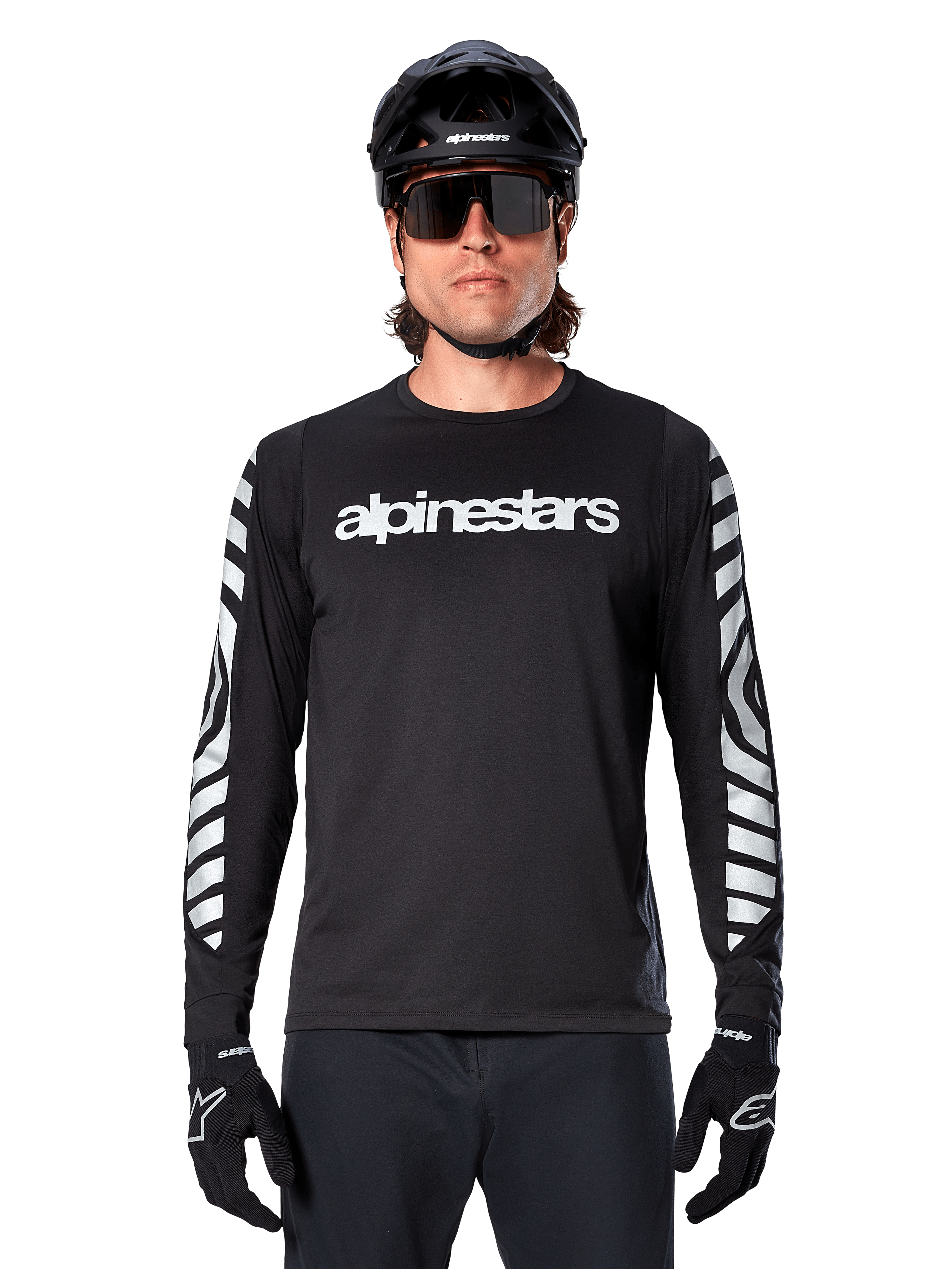 Mannelijke rijder in Alpinestars A-Dura Dri Oscar Jersey met lange mouwen, zwart reflecterend, voorzien van een wit Alpinestars-woordmerk op de borst en zilveren reflecterende graphics op de mouwen, gecombineerd met een zwarte MTB-helm, zonnebril en zwarte handschoenen, performance fietskleding