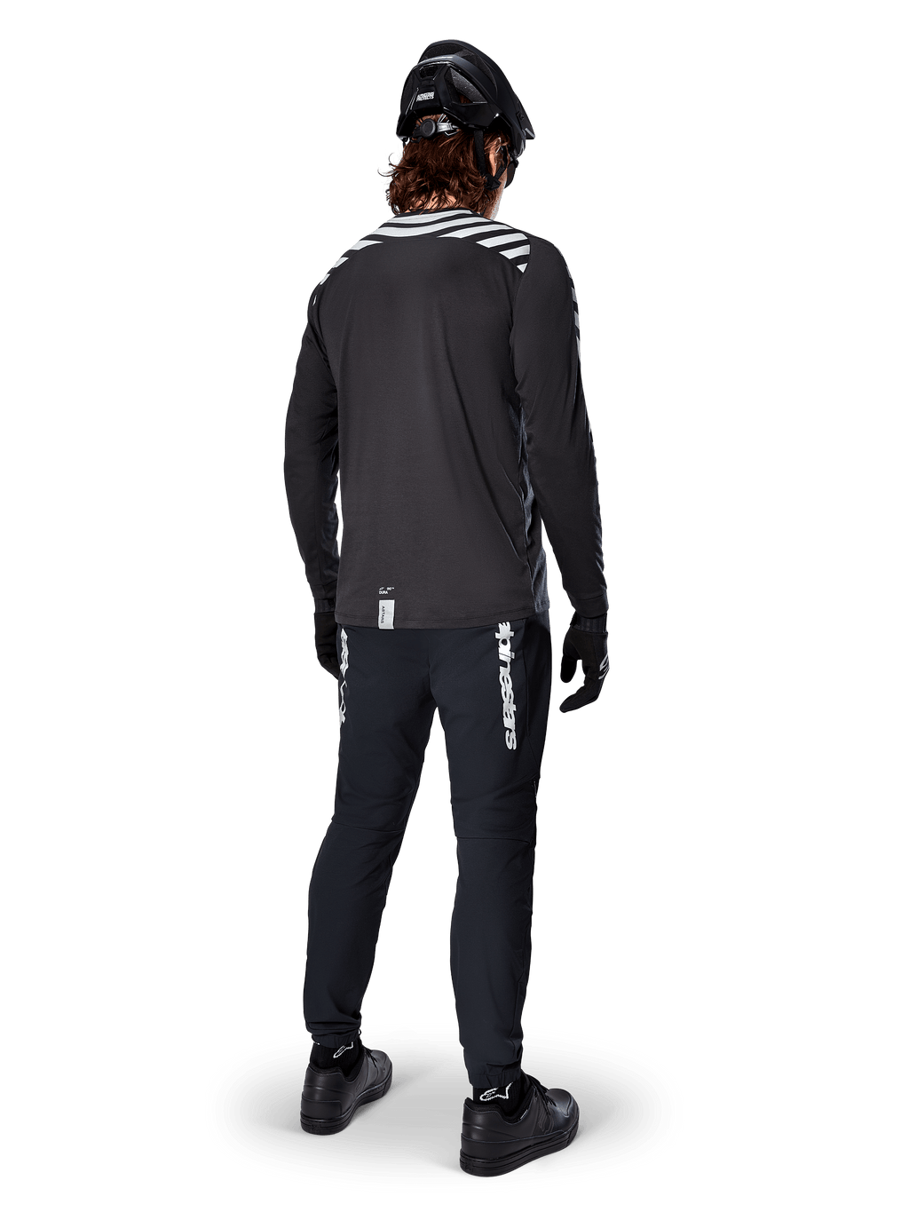 Mountainbiker die de Alpinestars A-Dura Dri Oscar Jersey met lange mouwen in Black Reflective draagt, van achteren gezien, gecombineerd met een zwarte technische broek, helm en handschoenen, voorzien van reflecterende streepgraphics op de schouders en een langere achterzijde voor off-road fietsen.