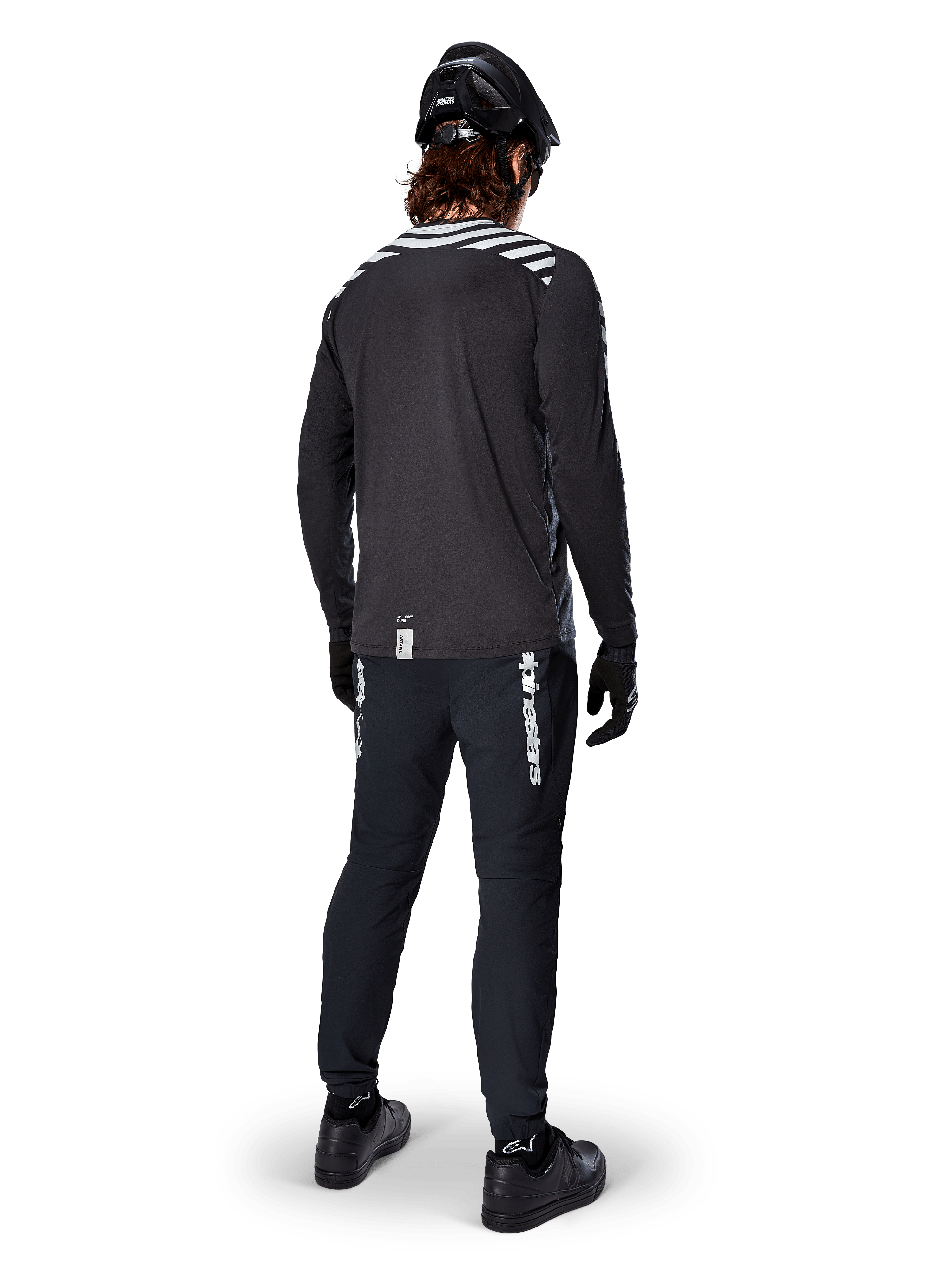Mountainbiker die de Alpinestars A-Dura Dri Oscar Jersey met lange mouwen in Black Reflective draagt, van achteren gezien, gecombineerd met een zwarte technische broek, helm en handschoenen, voorzien van reflecterende streepgraphics op de schouders en een langere achterzijde voor off-road fietsen.