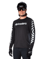 Alpinestars A-Dura Dri Oscar fietsshirt met lange mouwen, MTB-fietsshirt, zwart reflecterend, met een wit woordmerk op de borst en opvallende reflecterende cirkelvormige graphics op de mouwen, gedragen met een zwarte helm en zonnebril, technische vochtafvoerende kleding voor off-road mountainbiken