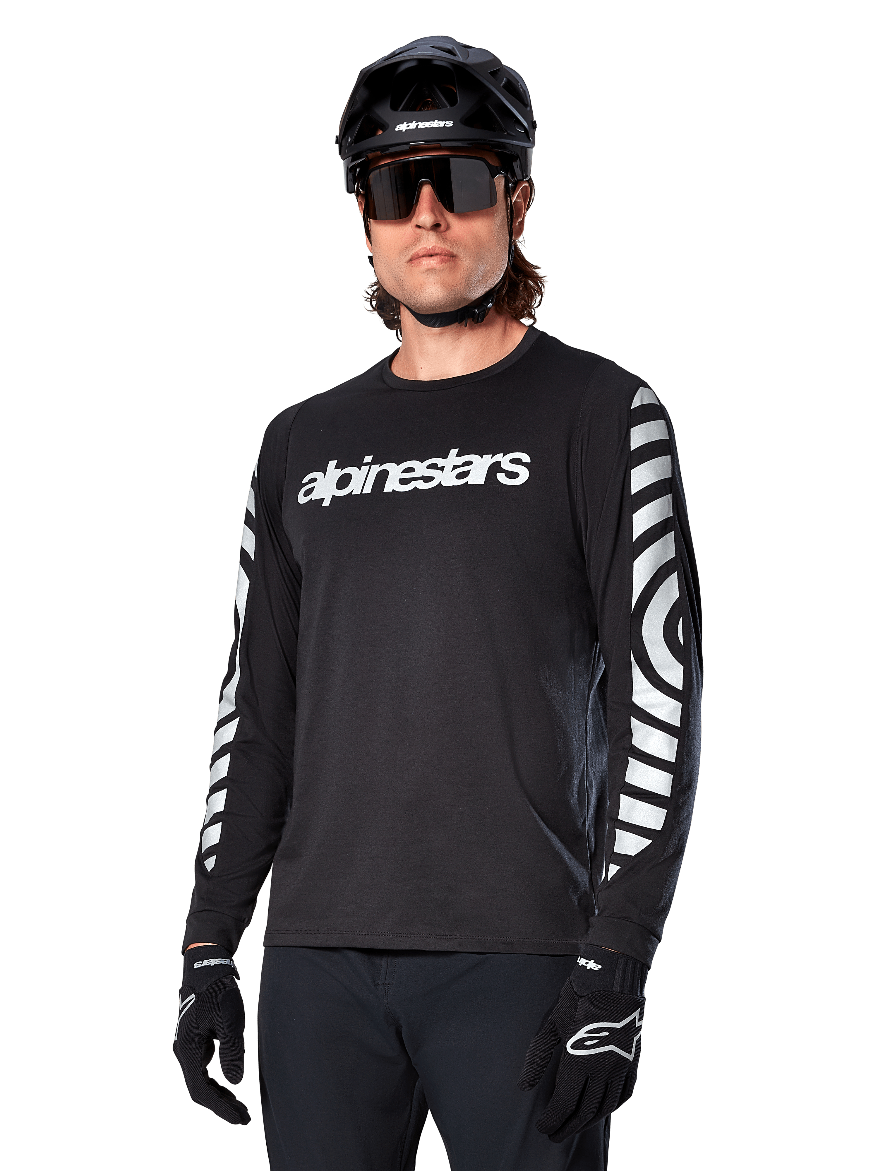 Alpinestars A-Dura Dri Oscar fietsshirt met lange mouwen, MTB-fietsshirt, zwart reflecterend, met een wit woordmerk op de borst en opvallende reflecterende cirkelvormige graphics op de mouwen, gedragen met een zwarte helm en zonnebril, technische vochtafvoerende kleding voor off-road mountainbiken