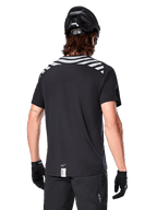Alpinestars A-Dura Dri Oscar Jersey, MTB-jersey met korte mouwen, Black Reflective, achteraanzicht op model met fietshelm en handschoenen, met reflecterende grijze schouderstrepen en een verlengd achterpand, vochtafvoerende technische stof voor mountainbiken