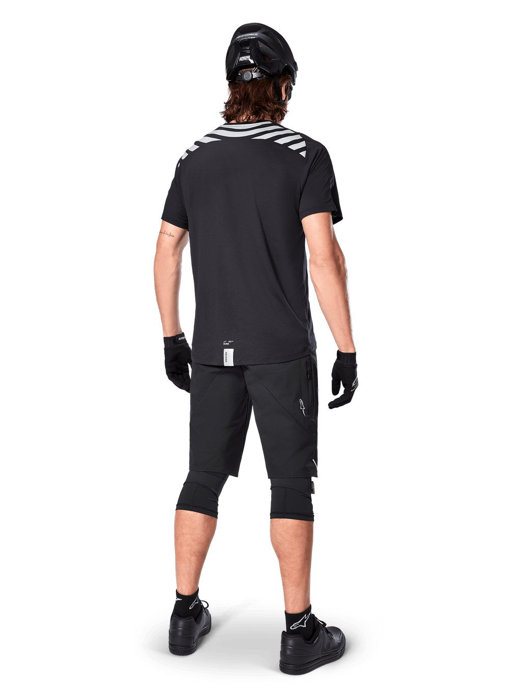 Alpinestars A-Dura Dri Oscar jersey met korte mouwen, Black Reflective, achteraanzicht van een rijder die de jersey draagt met een zwarte mountainbike short, helm en kniebeschermers, voorzien van reflecterende graphics op de schouders en een verlengd achterpand voor MTB-rijden.