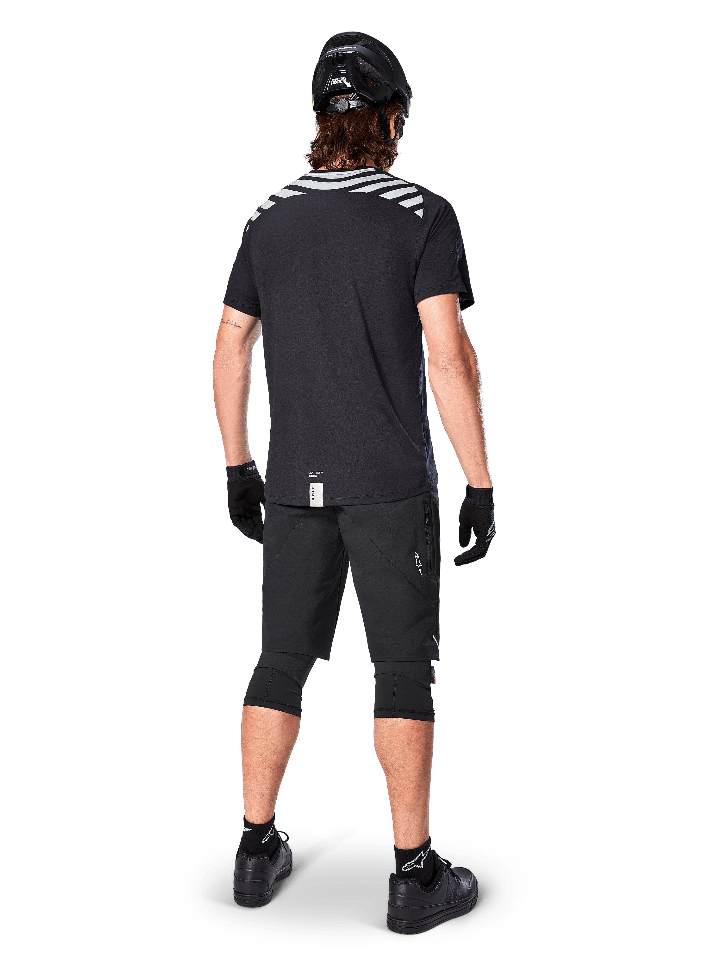 Alpinestars A-Dura Dri Oscar jersey met korte mouwen, Black Reflective, achteraanzicht van een rijder die de jersey draagt met een zwarte mountainbike short, helm en kniebeschermers, voorzien van reflecterende graphics op de schouders en een verlengd achterpand voor MTB-rijden.