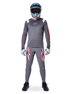 MTB rider wearing Alpinestars A-Supra Astar Long Sleeve Jersey, Dark Gray with red accents and white chest logo, matching gray pants with large red star graphics, blue full-face helmet with goggles, light gray gloves, and black shoes, professional mountain bike gear ensemble