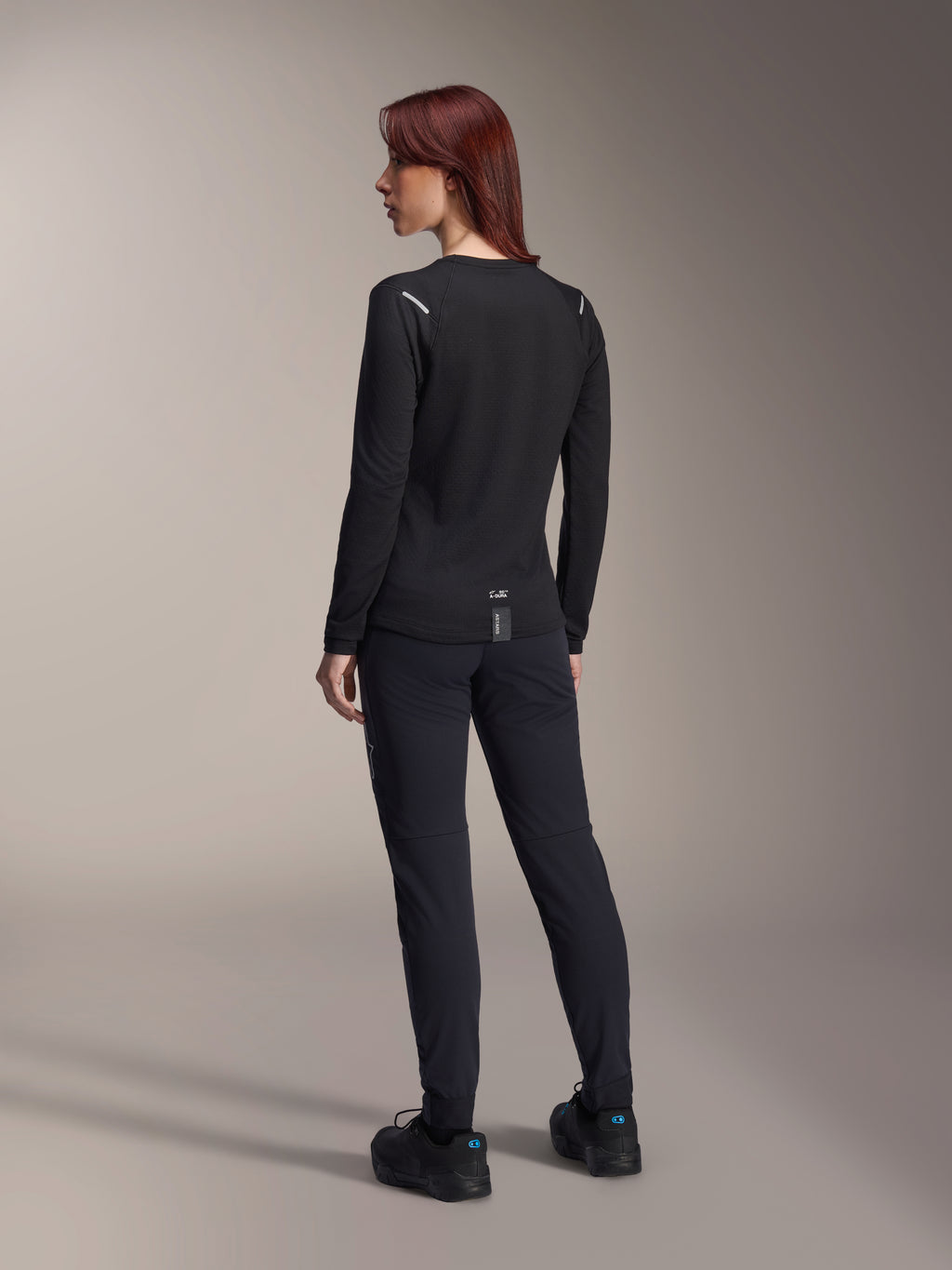 Alpinestars Stella A-Dura Thermal Jersey voor dames, MTB-shirt met lange mouwen in het zwart, achteraanzicht met reflecterende schouderaccenten en verlengde drop tail, gedragen met zwarte broek en schoenen, performance kleding voor mountainbiken