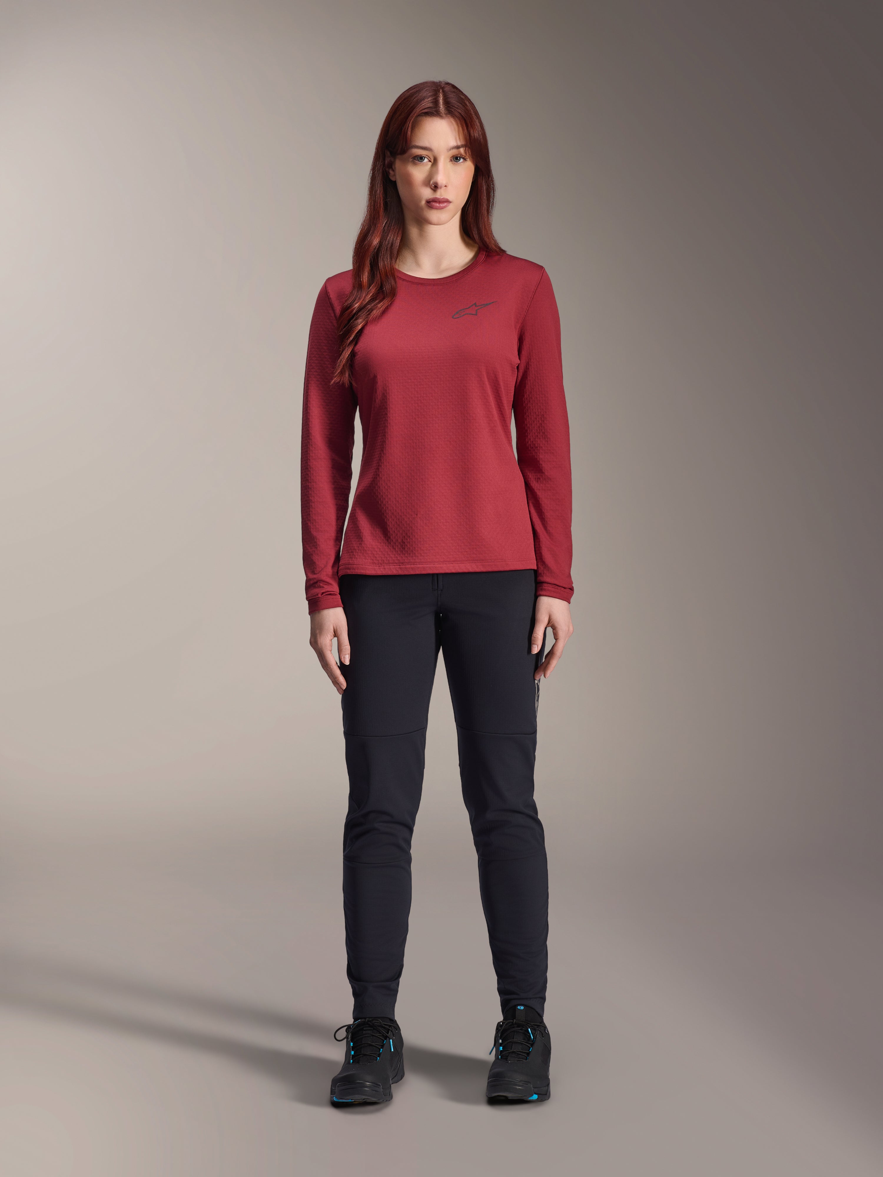 Alpinestars Stella A-Dura Thermal Jersey voor dames, fietsshirt met lange mouwen, bordeauxrood, gedragen door vrouwelijk model met zwarte broek en zwarte schoenen, subtiel logo op de borst, reguliere pasvorm voor mountainbiken