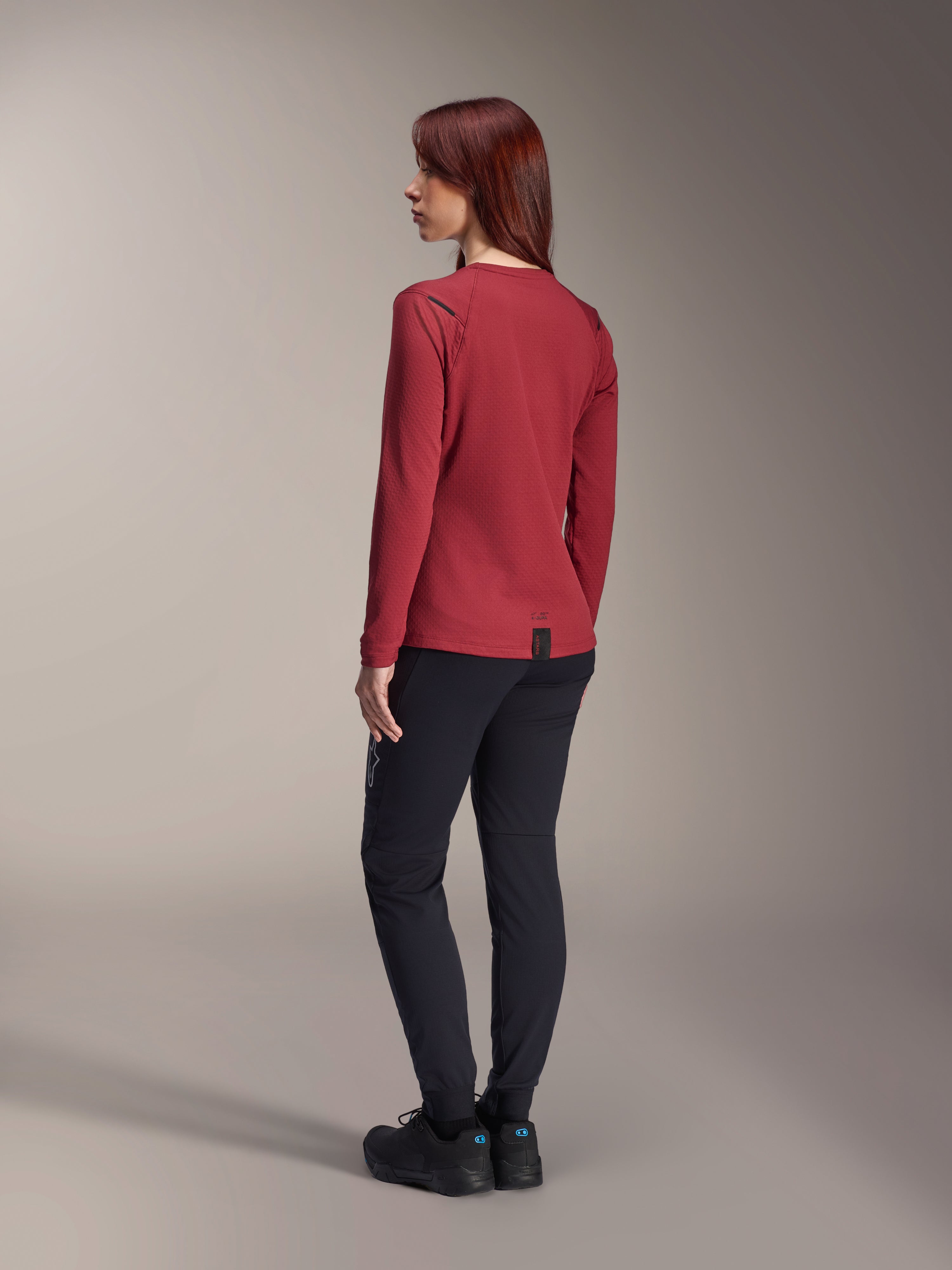 Alpinestars Stella A-Dura Thermal Jersey, mountainbike-jersey voor dames met lange mouwen, bordeauxrood, achteraanzicht met de nadruk op de verlengde achterkant en de textuur van de thermische stof, gedragen met een zwarte MTB-broek en zwart schoeisel