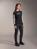 Vrouw in Alpinestars MTB-kleding, zwarte Stella A-Dura Elite Jersey met wit logo op de borst en witte panelen op de mouwen, zwarte Stella A-Dura broek, zwarte fietsschoenen met blauwe accenten, volledige performance outfit voor mountainbiken