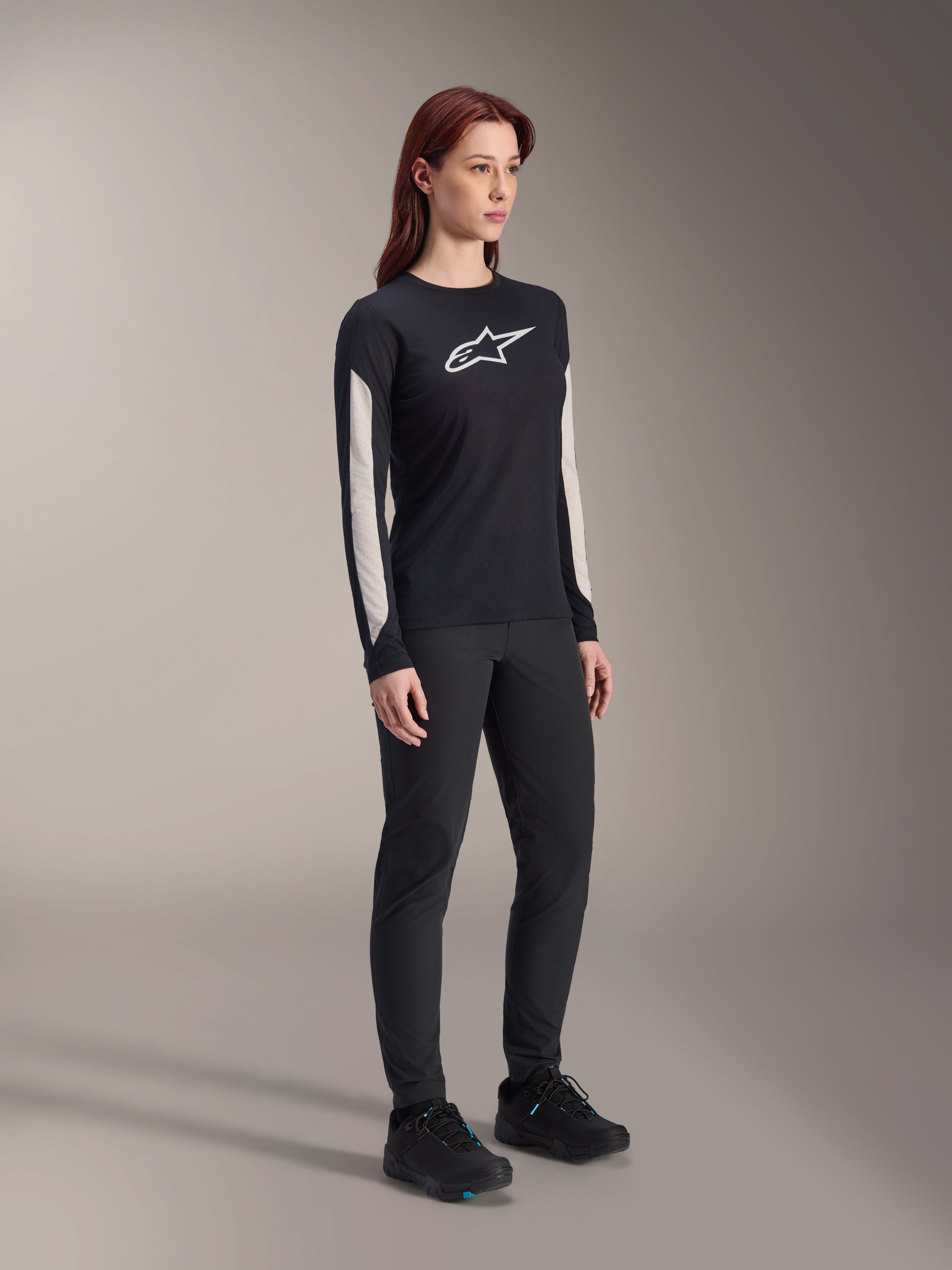 Vrouw in Alpinestars MTB-kleding, zwarte Stella A-Dura Elite Jersey met wit logo op de borst en witte panelen op de mouwen, zwarte Stella A-Dura broek, zwarte fietsschoenen met blauwe accenten, volledige performance outfit voor mountainbiken