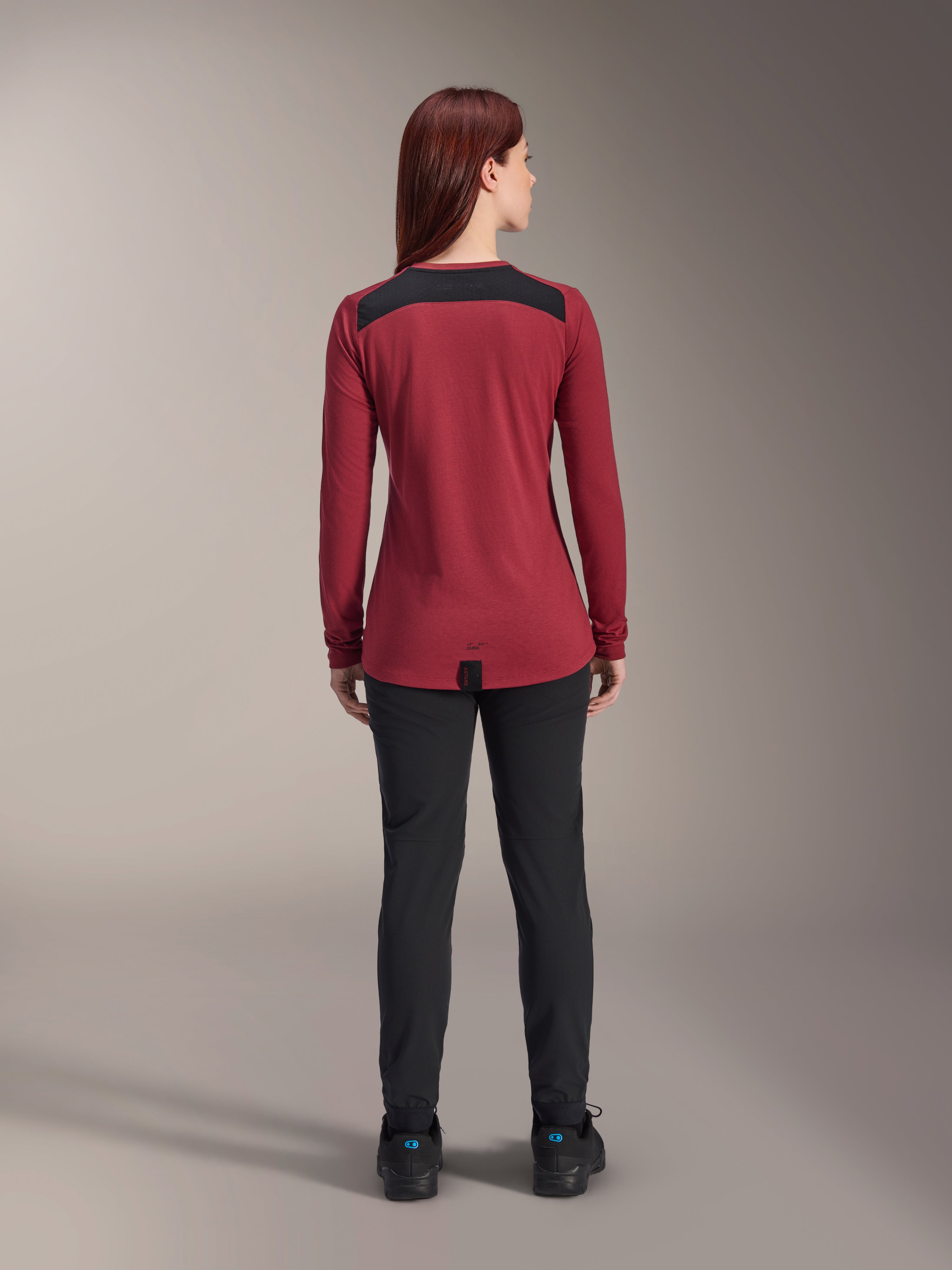 Alpinestars Stella A-Dura Dri Astar Long Sleeve Jersey voor dames, bordeauxrood met zwarte mesh schouderpanelen, technische MTB-kleding, van achteren afgebeeld met een zwarte broek, verlengde achterkant voor extra dekking tijdens het fietsen