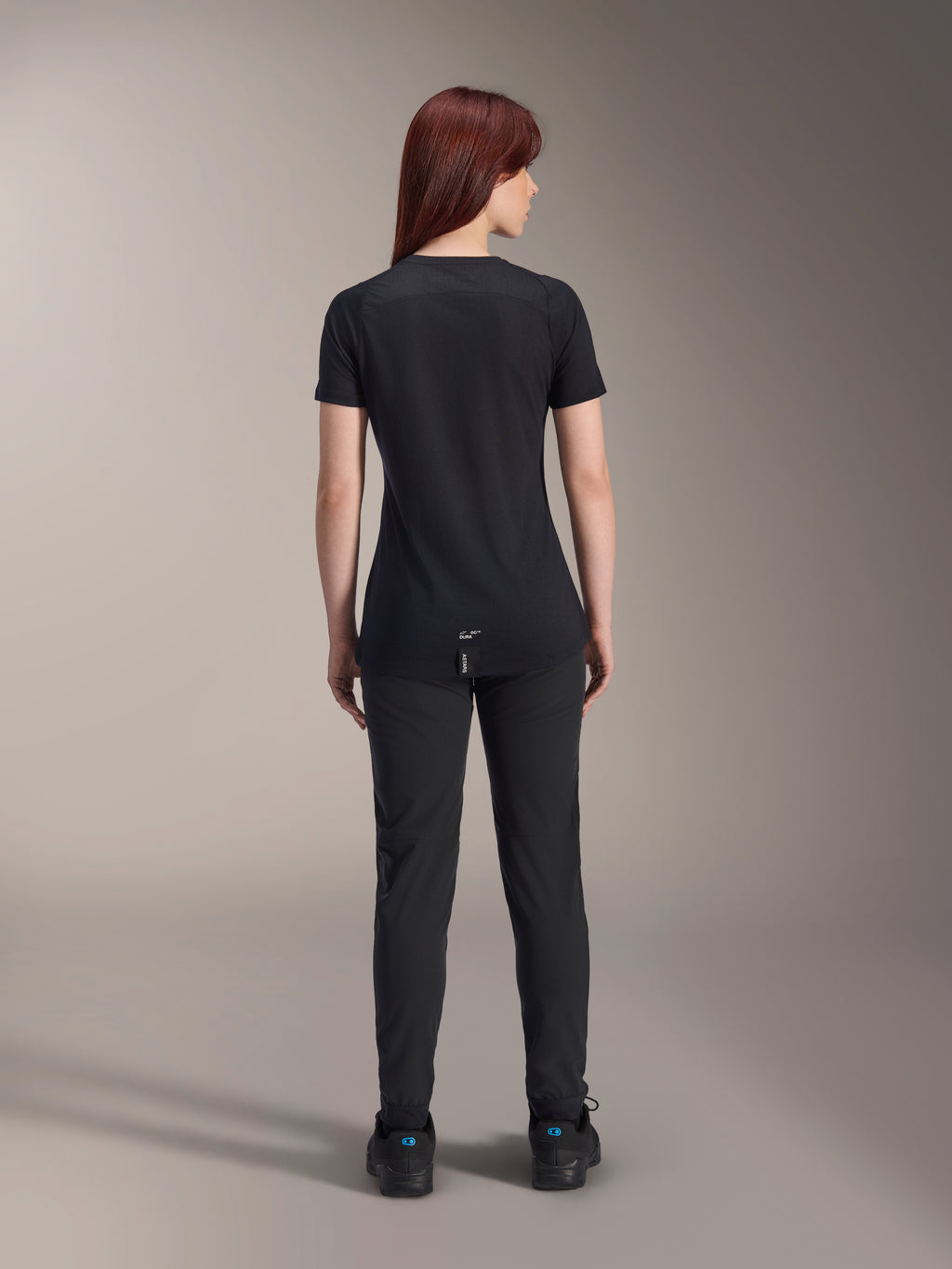 Alpinestars Stella A-Dura Dri Switch Jersey voor dames met korte mouwen, zwart, achteraanzicht op een model, voorzien van een verlengde achterkant en technische mesh-panelen, gecombineerd met een zwarte broek, performance MTB-wielerkleding