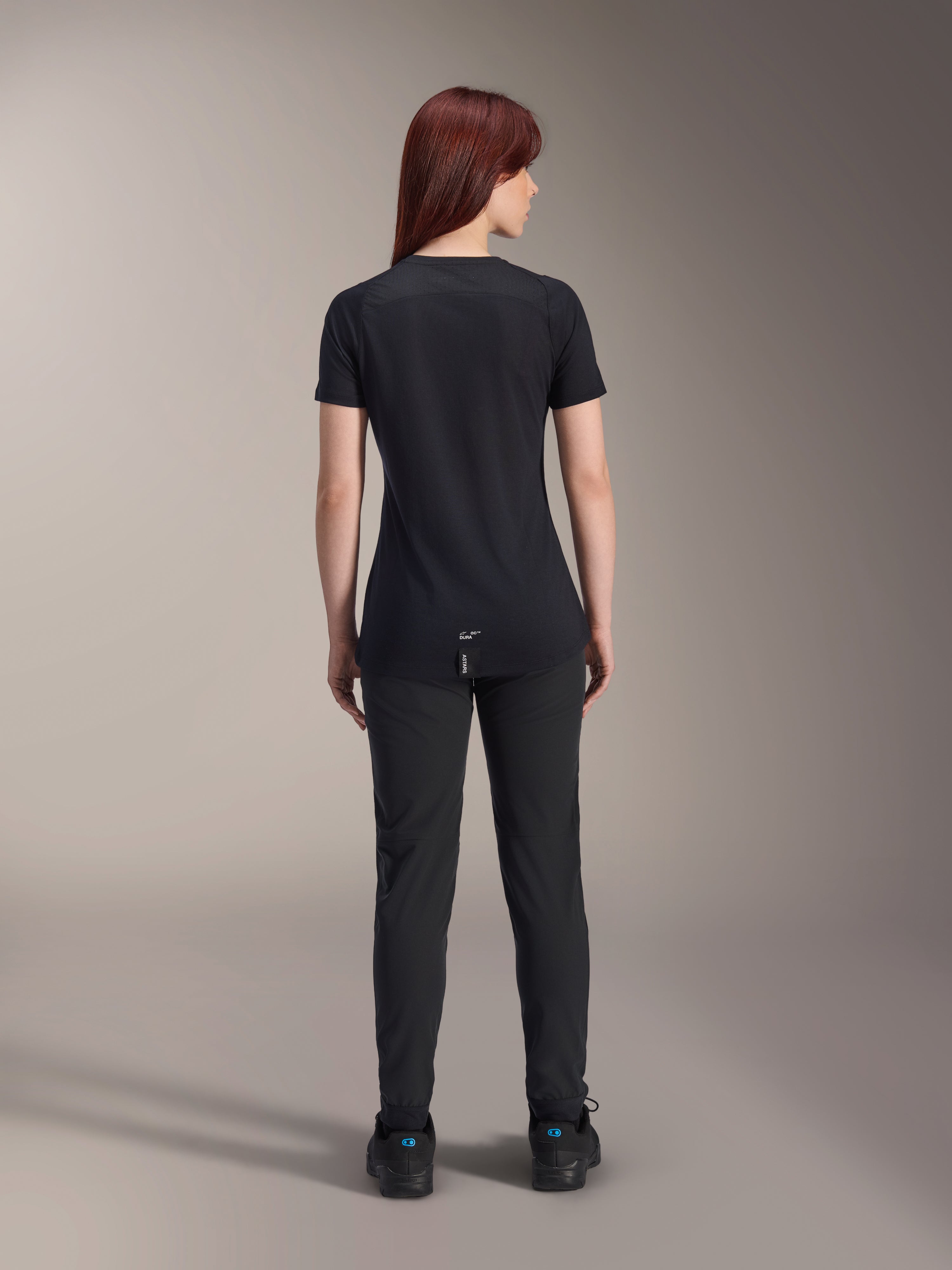 Alpinestars Stella A-Dura Dri Switch Jersey voor dames met korte mouwen, zwart, achteraanzicht op een model, voorzien van een verlengde achterkant en technische mesh-panelen, gecombineerd met een zwarte broek, performance MTB-wielerkleding