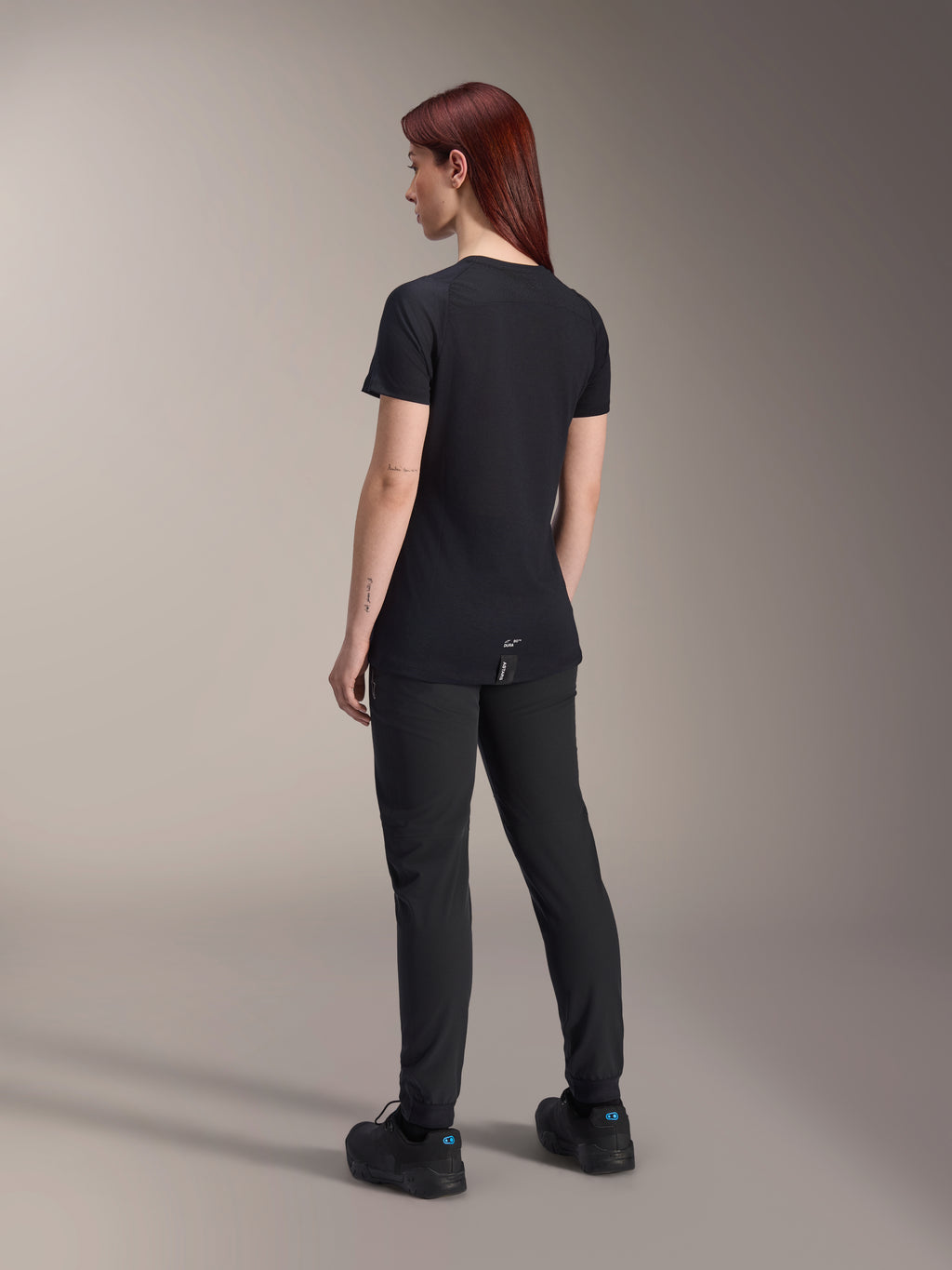 Alpinestars Stella A-Dura Dri Switch jersey met korte mouwen in het zwart, dames mountainbike shirt, achteraanzicht, gecombineerd met zwarte MTB-broek en schoenen, technische pasvorm met verlengde achterkant voor extra bedekking, model met rood haar