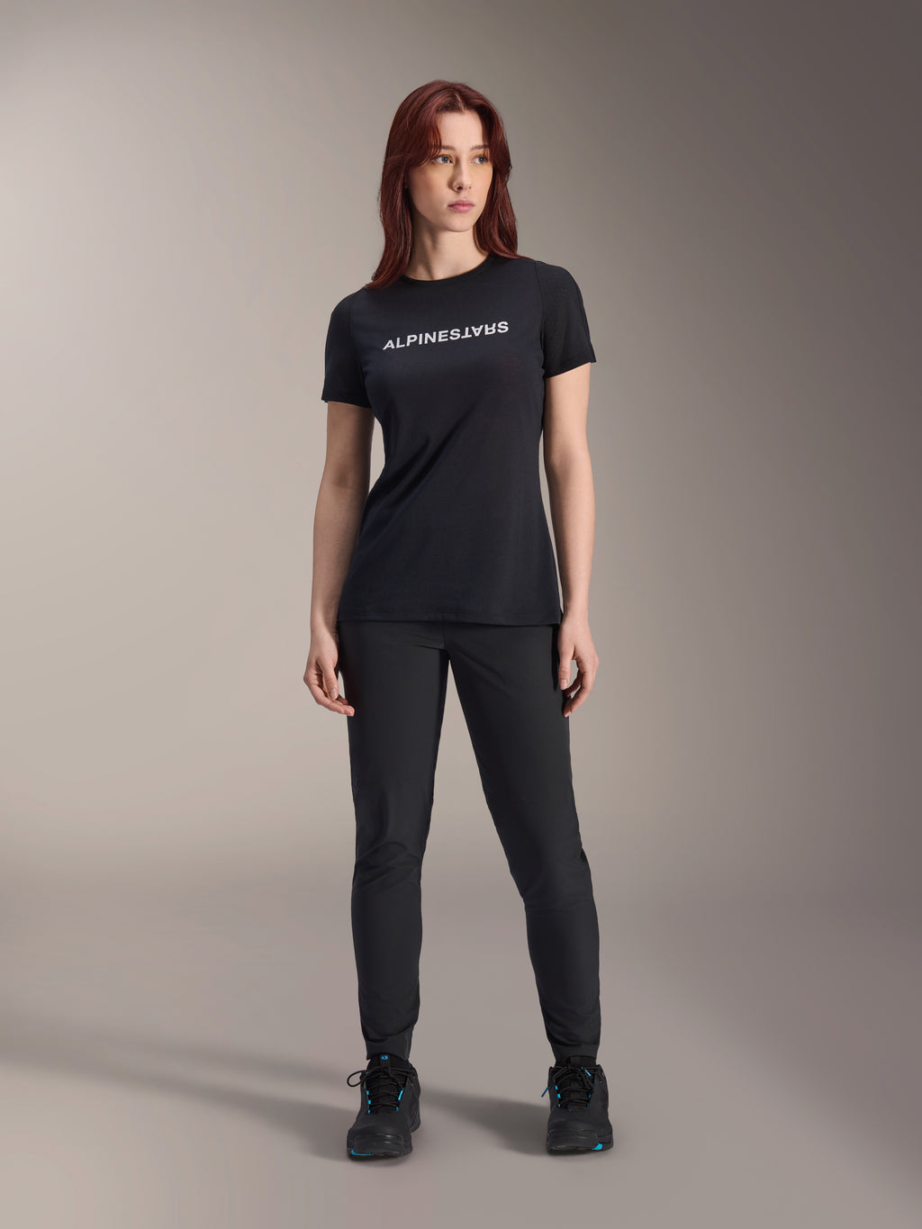 Model draagt Alpinestars Stella A-Dura Dri Switch jersey voor dames met korte mouwen in het zwart, met een wit logo op de borst, gecombineerd met een zwarte technische broek en zwarte sportschoenen, studioportret ten voeten uit