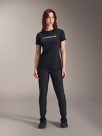 Model draagt Alpinestars Stella A-Dura Dri Switch jersey voor dames met korte mouwen in het zwart, met een wit logo op de borst, gecombineerd met een zwarte technische broek en zwarte sportschoenen, studioportret ten voeten uit