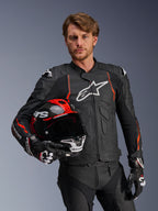 Alpinestars Dusk Airflow Leren Jas, zwart met Red Fluo accenten, gedragen door een mannelijk model die een rode en zwarte motorhelm vasthoudt, voorzien van geperforeerd leer voor maximale ventilatie en witte Alpinestars-branding, sportieve motorrijuitrusting