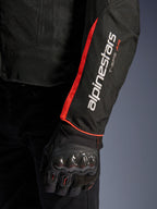 Alpinestars T-Sps Air V2 Jas, close-up van de mouw in Black Red Fluo, voorzien van witte Alpinestars en T-SPS AIR-logo's, felrode fluorescerende streep, gedragen met een zwarte beschermende motorhandschoen met carbonfiber knokkelbescherming
