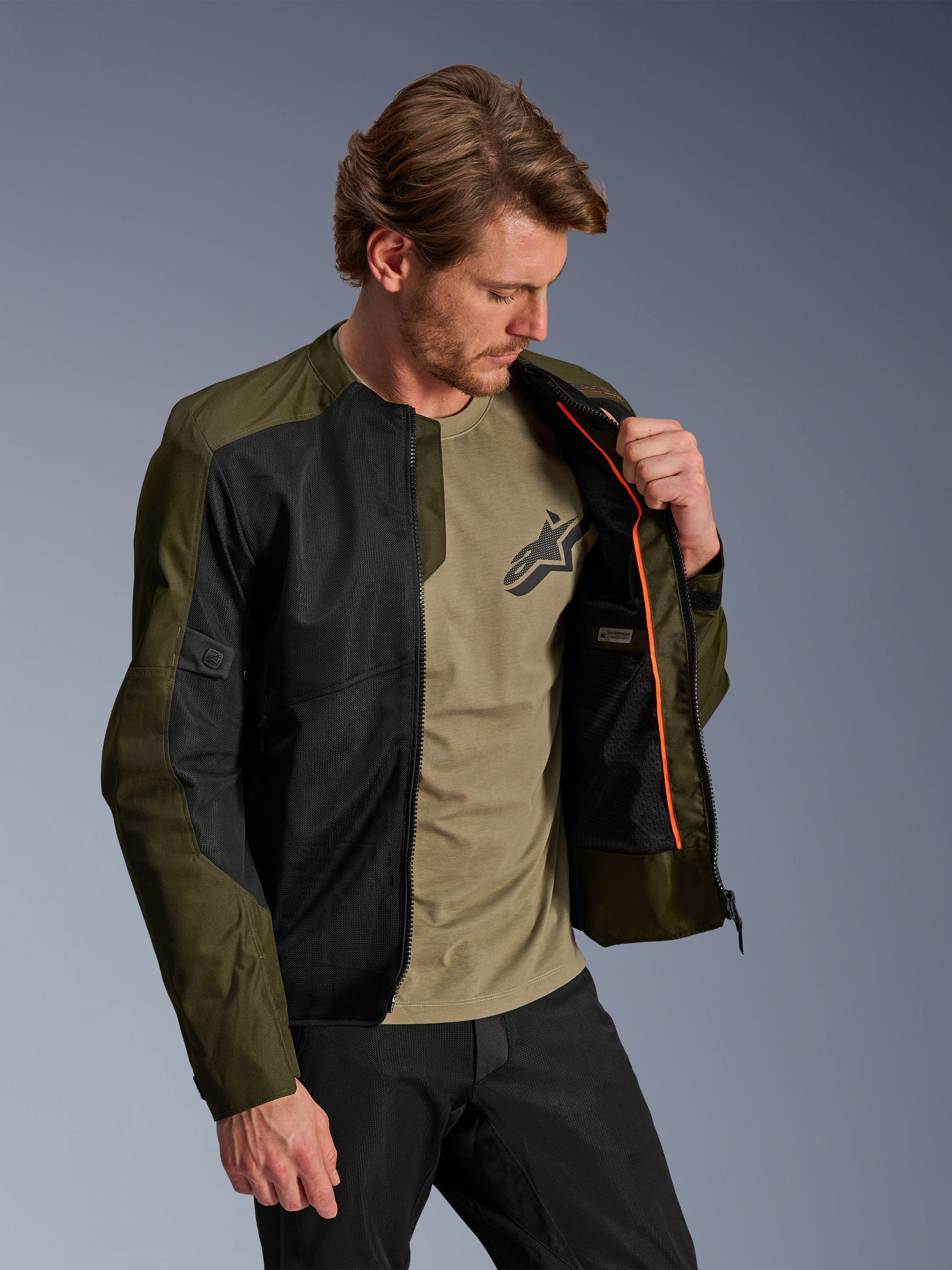 Alpinestars C-1 Air Jacket in Khaki en zwart, gedragen door een mannelijk model om de oranje binnenvoering en het waterdichte compartiment te tonen, voorzien van grote tech mesh-panelen voor luchtstroom, urban eigentijdse motorstijl gecombineerd met een bijpassend olijfgroen t-shirt en zwarte broek