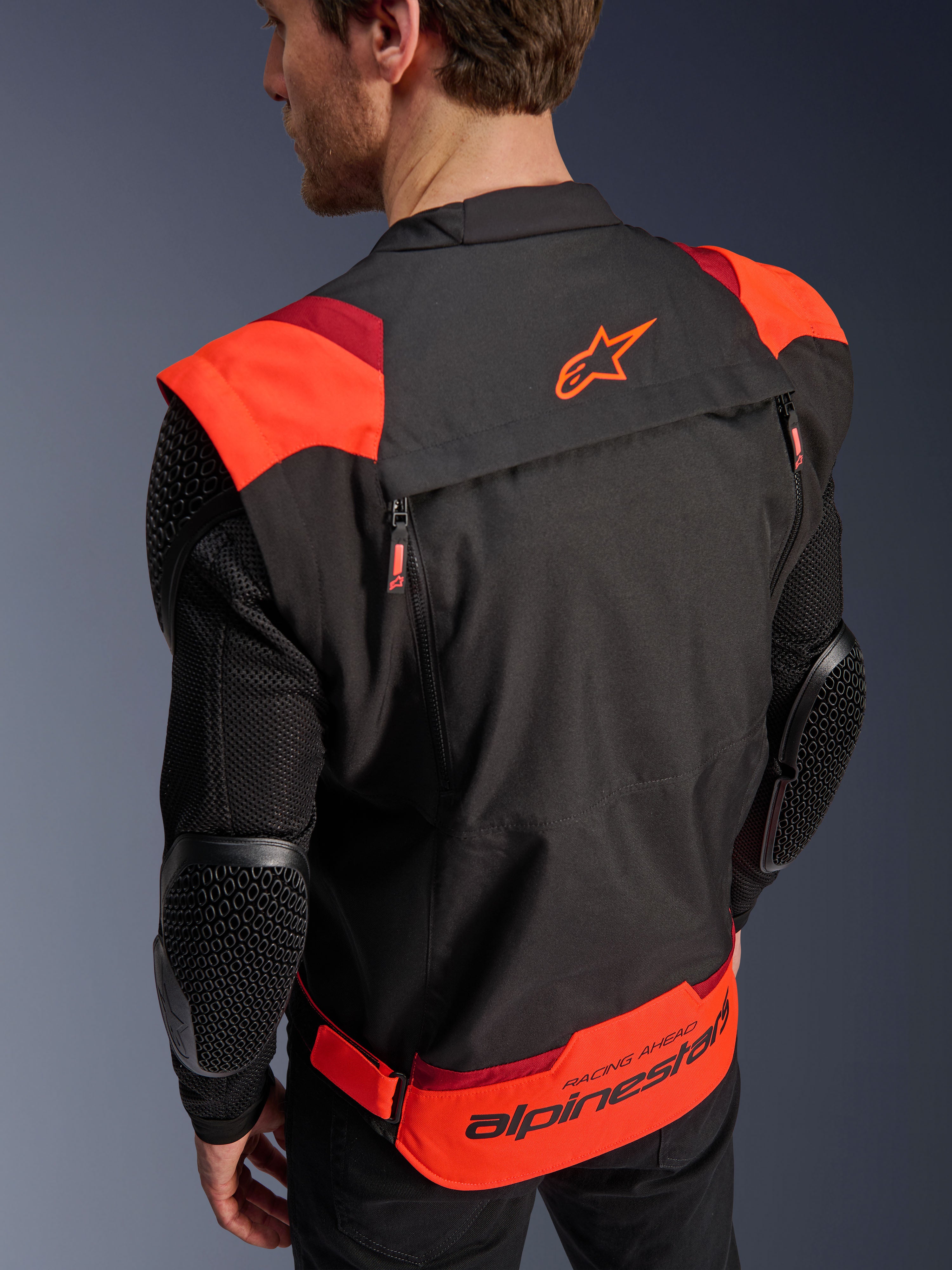Alpinestars T-Stunt Air jas, zwart, fluorood en donkerrood, achteraanzicht op een model, toont mesh mouwen met elleboogbeschermers met honingraatstructuur, Alpinestars-sterlogo op de bovenrug en opvallende branding op de onderrug met 'RACING AHEAD' tekst, modulair sportief ontwerp voor motorrijders.