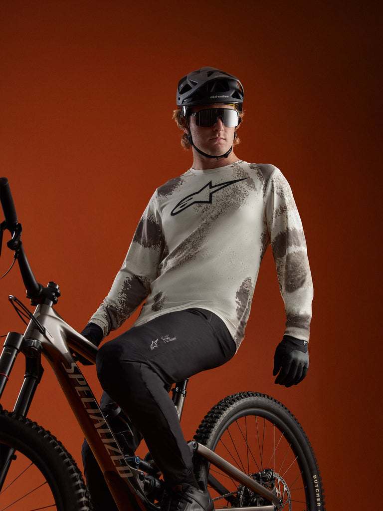 MTB-rijder in een A-Dura Ride Sand Camo jersey met lange mouwen en Alpinestars-logo op een mountainbike.