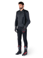 Alpinestars Missile V3 Leren Jas, zwart, gedragen door een rijder met een zwarte broek en zwart-rode racehandschoenen en laarzen, voorzien van geperforeerde leren panelen en strakke monochrome branding, professionele racing sport esthetiek