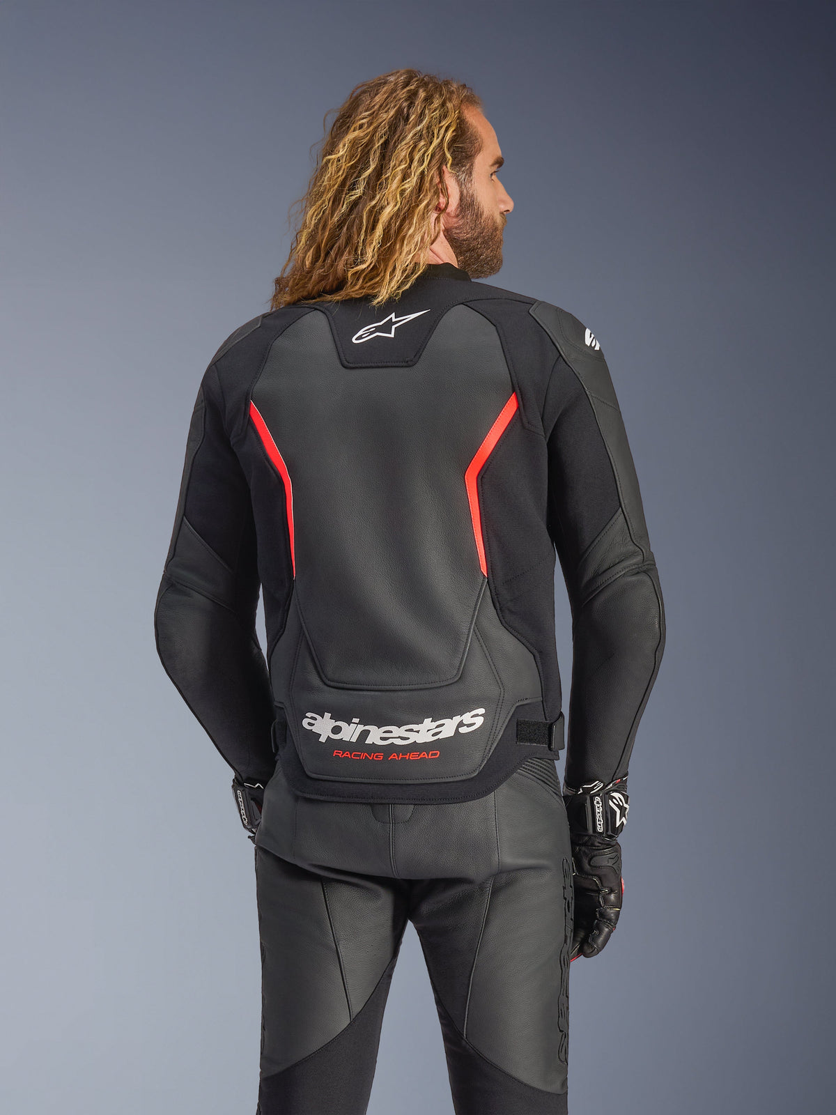 Alpinestars GP Force V2 Leren Jas, zwart en fluo rood, achteraanzicht op een model, met wit Alpinestars-logo en 'RACING AHEAD'-tekst, gecombineerd met een zwarte leren motorbroek en handschoenen