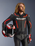 Alpinestars GP Force V2 leren jas, zwart en rood fluo, gedragen door een mannelijk model met een bijpassende helm, voorzien van witte Alpinestars-logo's en rode accenten, gecombineerd met een leren broek en racehandschoenen, sportieve motoruitrusting voor op de weg en het circuit.