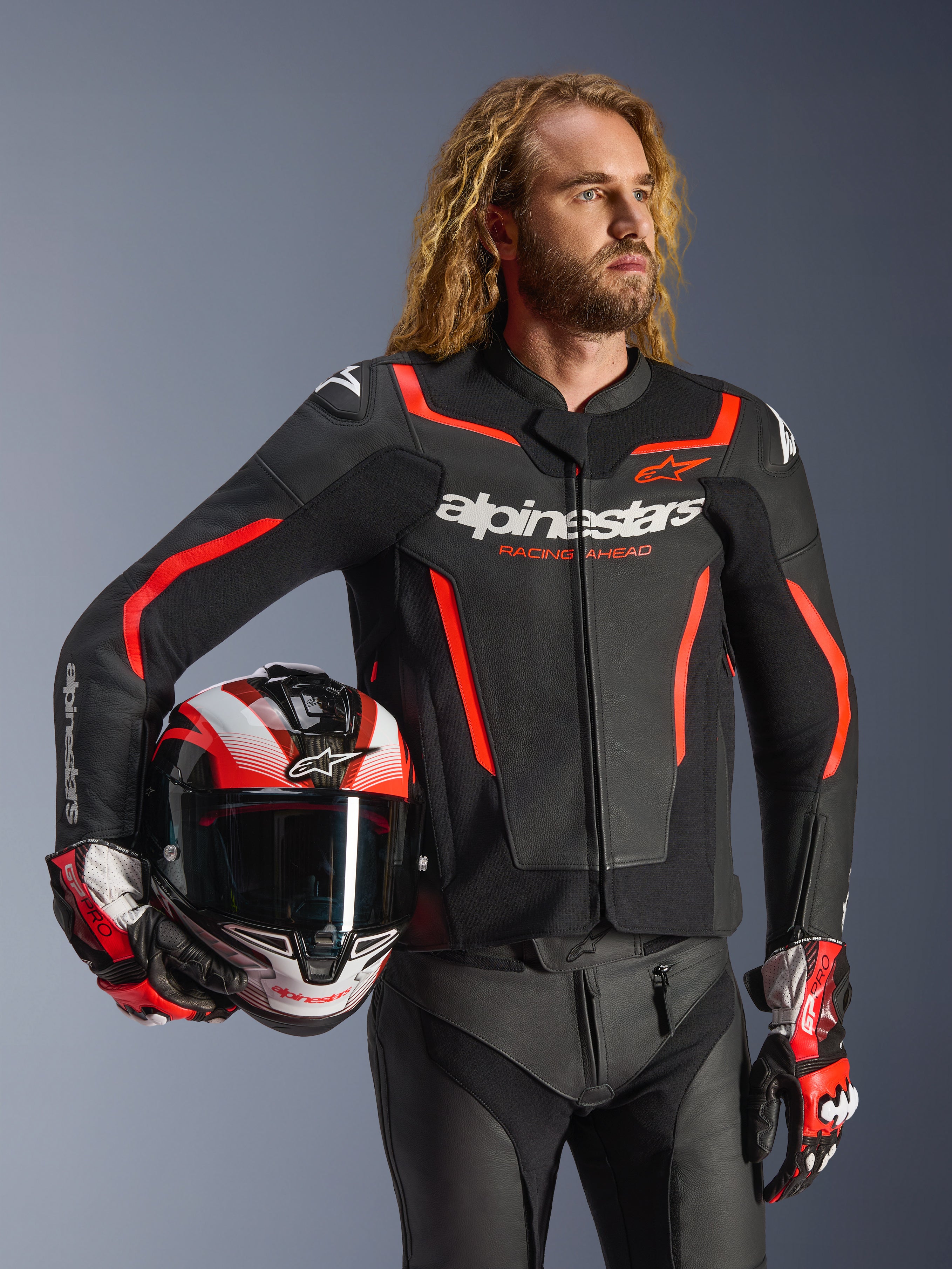 Alpinestars GP Force V2 leren jas, zwart en rood fluo, gedragen door een mannelijk model met een bijpassende helm, voorzien van witte Alpinestars-logo's en rode accenten, gecombineerd met een leren broek en racehandschoenen, sportieve motoruitrusting voor op de weg en het circuit.