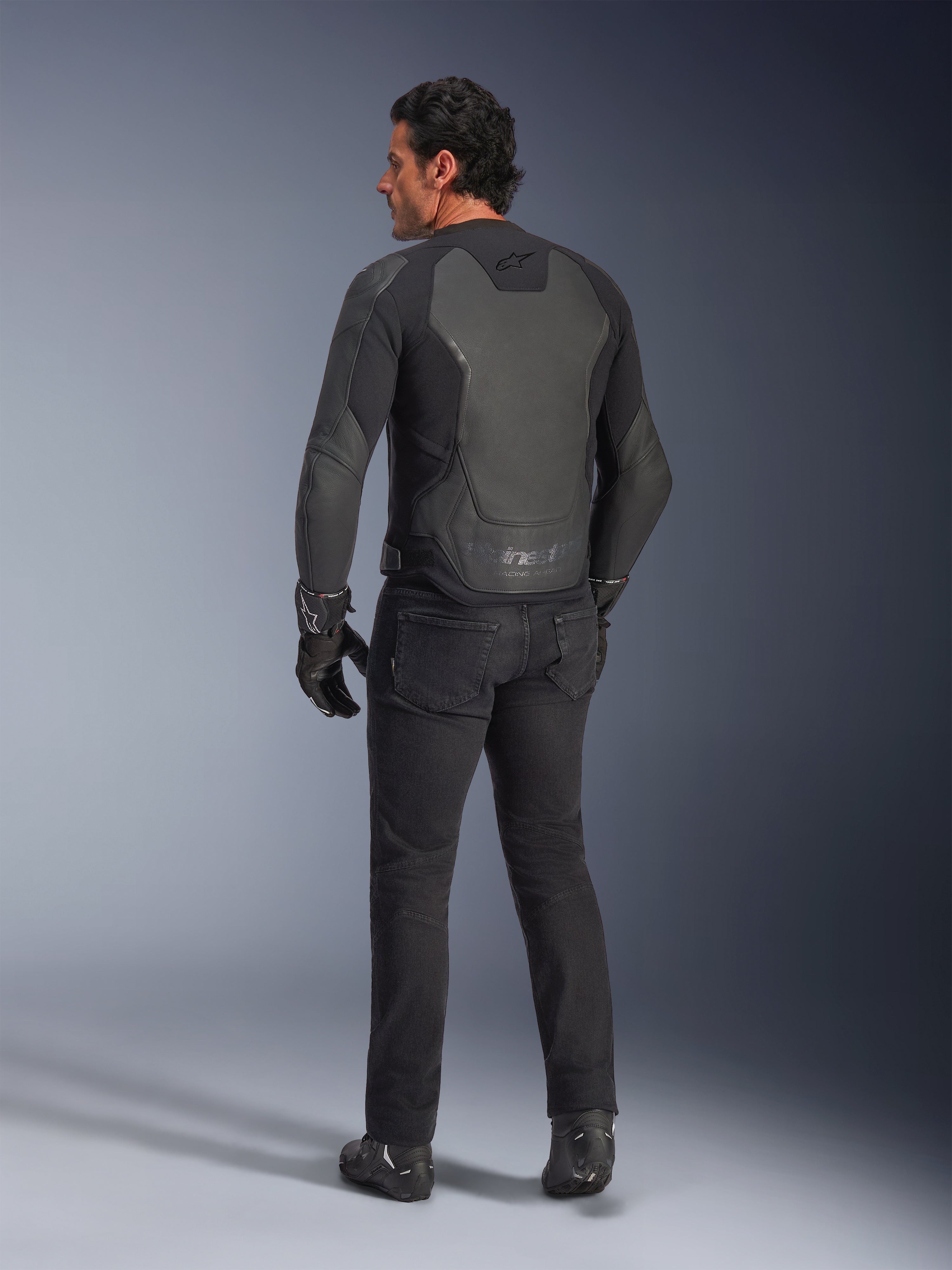 Achteraanzicht van een model die de Alpinestars GP Force V2 leren jas draagt in Black Black, voorzien van een meerpanelige leren constructie en sportieve pasvorm, gecombineerd met een zwarte motorbroek, handschoenen en laarzen voor een complete, op motorracen geïnspireerde look.