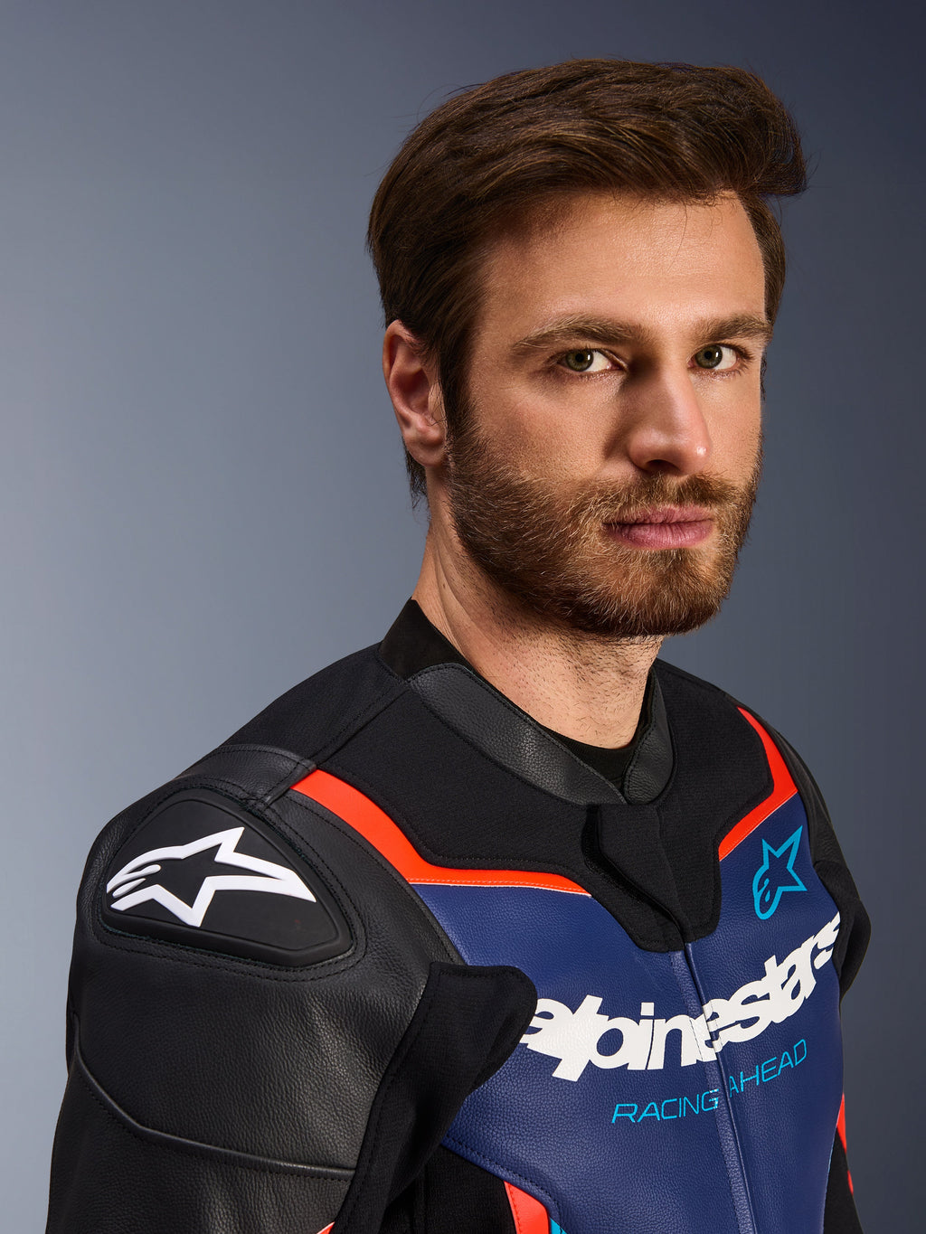 Alpinestars GP Force V2 Leather Jacket, Black Blue and Red Fluo, close-up portrait of a male rider, featuring white Alpinestars logo on black DFS shoulder shields, blue leather chest panel with white branding and 'Racing Ahead' text, red fluo accents, premium sport motorcycle gear.