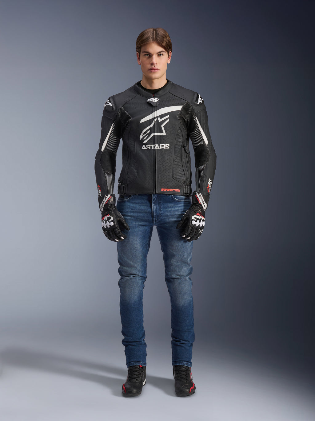 Alpinestars GP Plus R V4 Rideknit leren jas in zwart en wit, gedragen door een mannelijk model met blauwe motorjeans, zwarte motorhandschoenen en zwarte motorschoenen, voorzien van een groot wit Astars-logo op de borst, geperforeerde leren panelen en witte racestrepen op de mouwen, staand vooraanzicht.