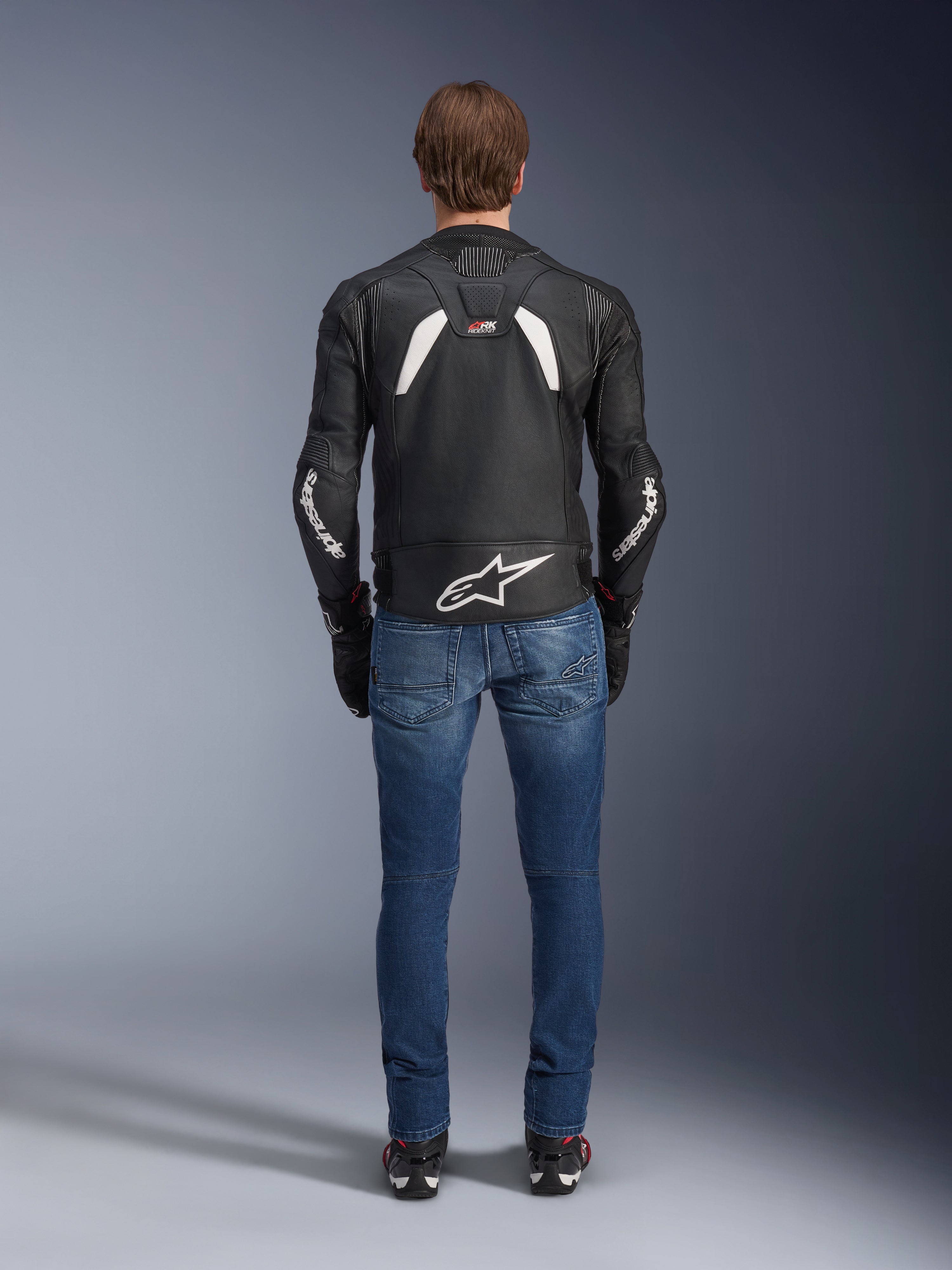 Achteraanzicht van de Alpinestars GP Plus R V4 Rideknit Leren Jas, zwart en wit, voorzien van een groot wit logo op de onderrug en branding op de mouwen, gedragen met een blauwe motorjeans en zwarte laarzen, wat de technische constructie en sportieve pasvorm benadrukt.