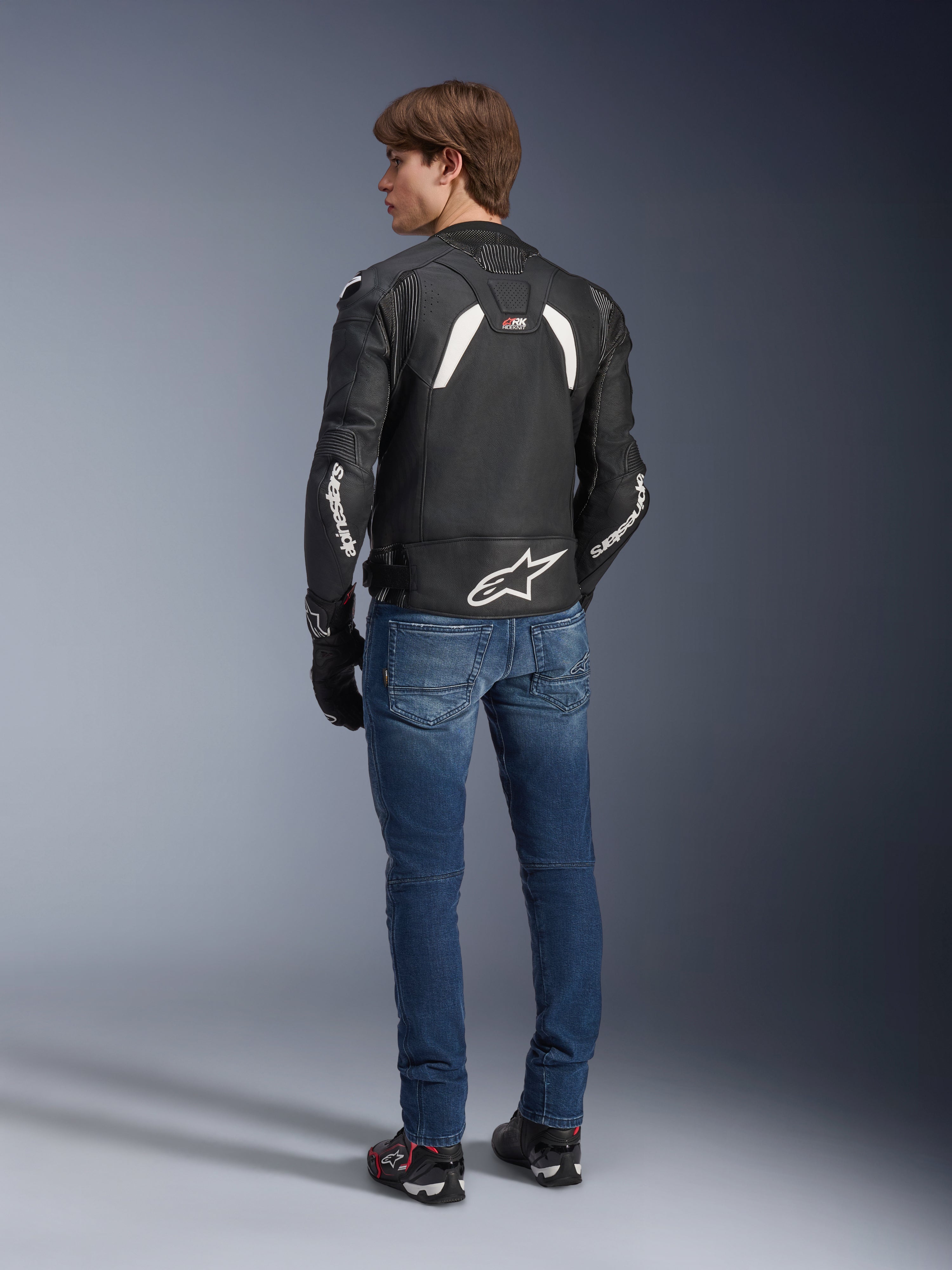 Alpinestars GP Plus R V4 Rideknit leren jas in zwart en wit, achteraanzicht op een model, met witte Alpinestars-branding op de mouwen en een groot sterlogo op de onderrug, gedragen met een blauwe jeans en zwarte motorschoenen, technische motoruitrusting.