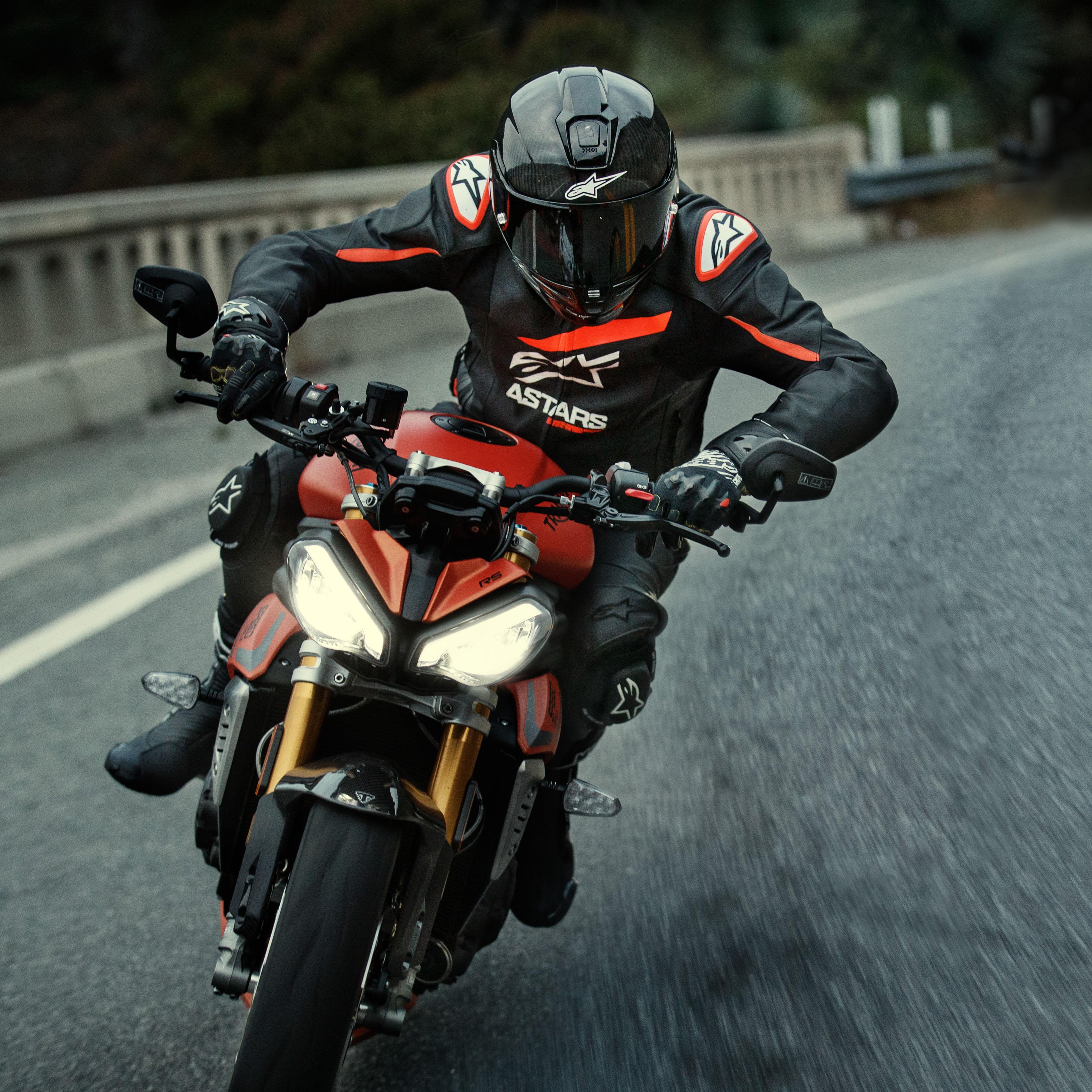 Motorrijder op een rode sportmotor, Alpinestars GP Plus R V4 Rideknit leren jas, zwart met Red Fluo en witte details, voorzien van wit Alpinestars-logo en 'ASTARS'-tekst, gecombineerd met zwarte racehelm en handschoenen, performance motorkleding in actie op de weg.