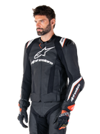 Mannelijk model in de Alpinestars Missile V3 Ignition leren jas, zwart met witte en oranje accenten, voorzien van opvallende witte logo's, geperforeerde leren delen en beschermende schouderprotectie, gestyled met racehandschoenen voor een sportieve race-look.